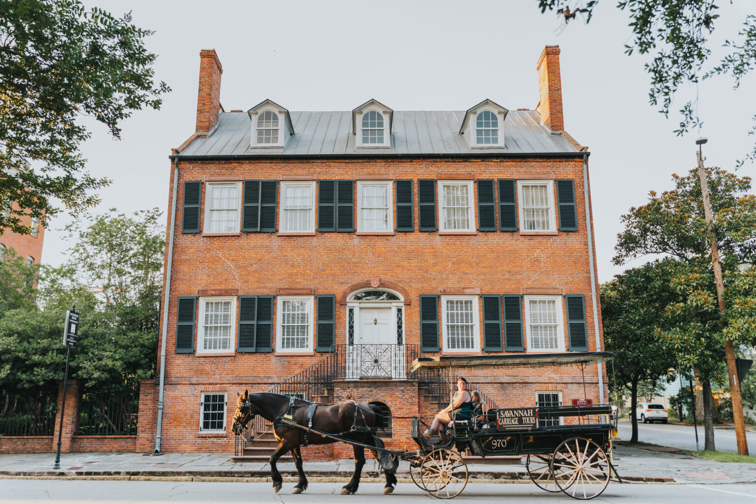 Elopement Wedding A horse-drawn carriage travels in front of a large, historic brick building with white trim and green shutters, perfect for romantic elopements. The building features three dormer windows on the roof, two chimneys, and a centered front door with a staircase leading up to it. Trees frame both sides of the scene. Elopements Inc