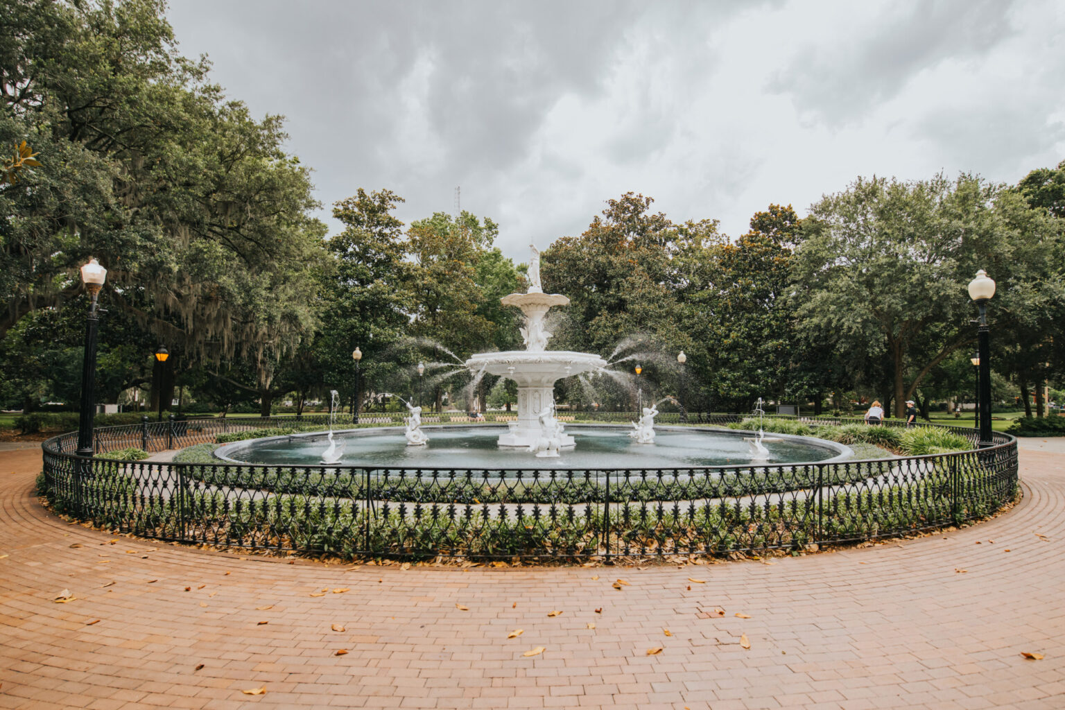 Elopement Wedding A round fountain with multiple tiers is in the center of a paved area surrounded by a black metal fence. Water streams elegantly from each tier. The area is encircled by lush green trees and lampposts, creating an idyllic spot for an elopement. The sky is overcast, suggesting an impending rainstorm. Elopements Inc