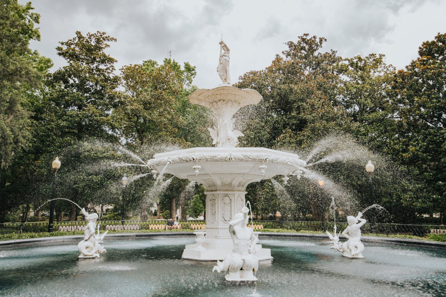 Elopement Wedding A large, ornate white fountain in a park is depicted, with statues of figures and water spouting from multiple points. The fountain, often chosen as a romantic spot for elopements, is set against a backdrop of lush green trees and a cloudy sky, creating a serene and picturesque scene. Elopements Inc