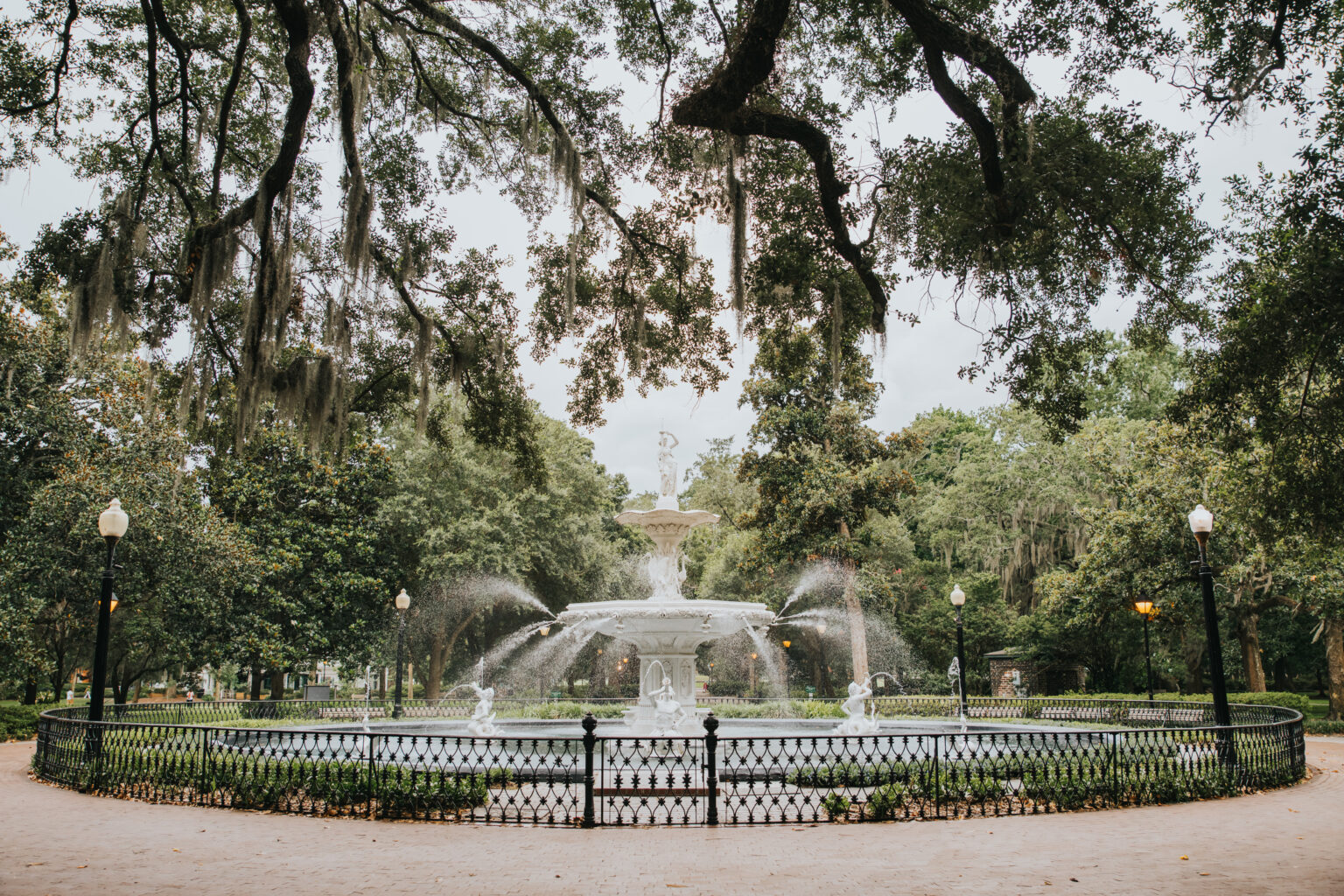 Elopement Wedding A large, ornate white fountain with multiple tiers of water spraying from it is set in the center of a lush, green park. Ideal for intimate elopements, the fountain is surrounded by a black wrought iron fence. Tall trees with hanging Spanish moss frame the scene, and lamp posts line the perimeter of the fountain area. Elopements Inc