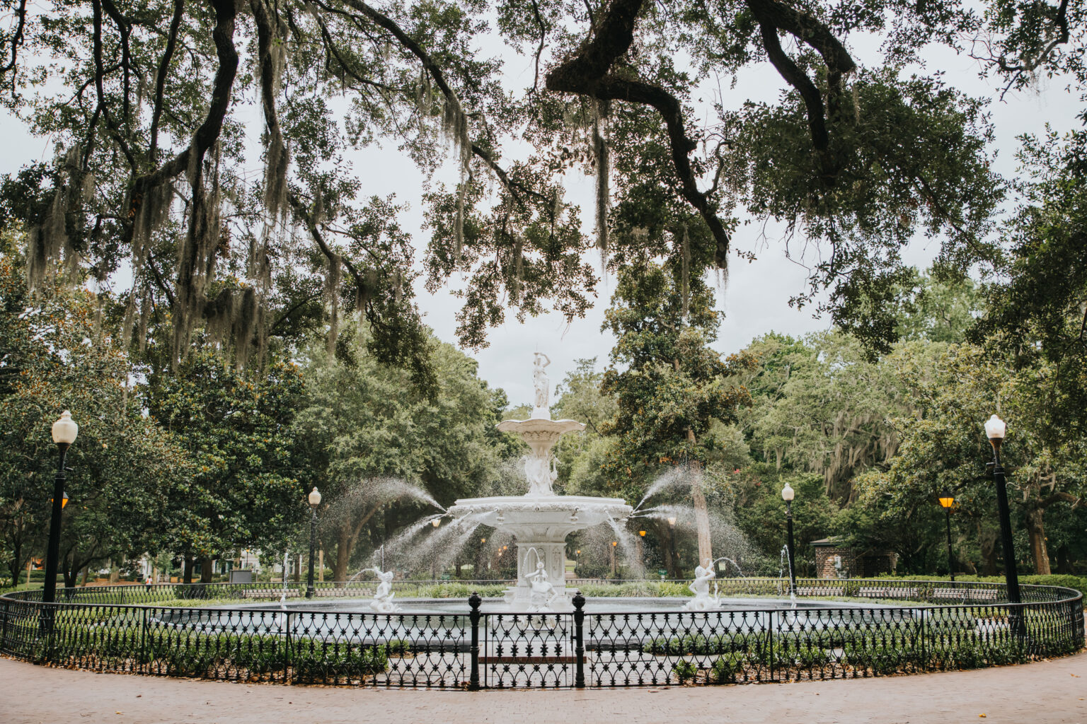 Elopement Wedding A large, ornate fountain is surrounded by wrought iron fencing in a lush, green park—perfect for an intimate elopement. Water cascades from the multi-tiered fountain, with lampposts evenly spaced around the perimeter. Tall, leafy trees with hanging moss frame the scene under a cloudy sky. Elopements Inc