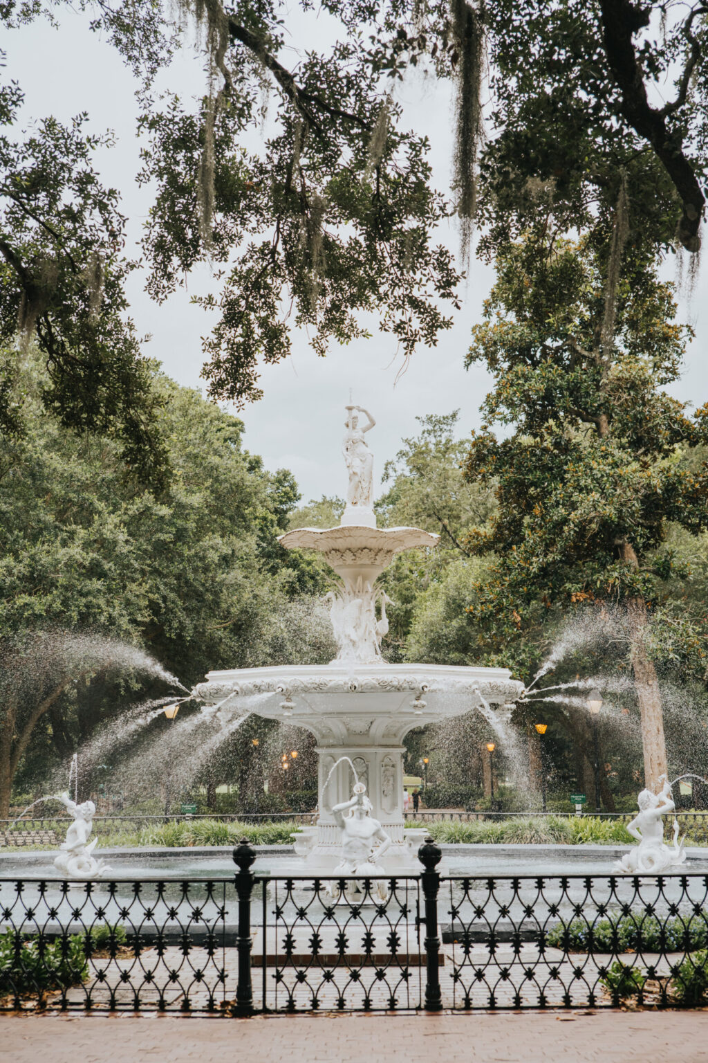 Elopement Wedding A large, ornate fountain stands in the center of a park, featuring multiple tiers and statues of figures and animals. Water streams from various points, creating a dynamic display. The fountain is surrounded by trees with hanging moss and a black wrought-iron fence—a perfect spot for intimate elopements. A paved path encircles the fountain. Elopements Inc