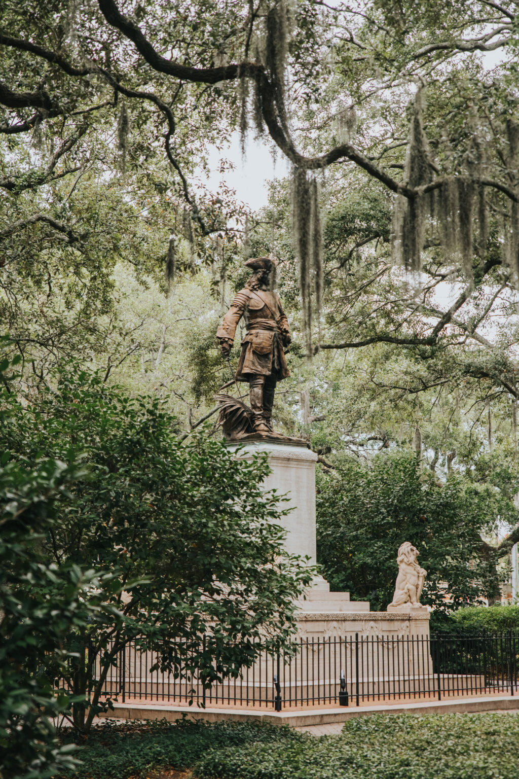 Elopement Wedding A historic statue stands atop a pedestal in a lush, green park where many couples dream of eloping. The figure is dressed in period clothing and is surrounded by trees draped with Spanish moss. A black iron fence encircles the statue, and a stone lion sculpture sits at the base. Elopements Inc