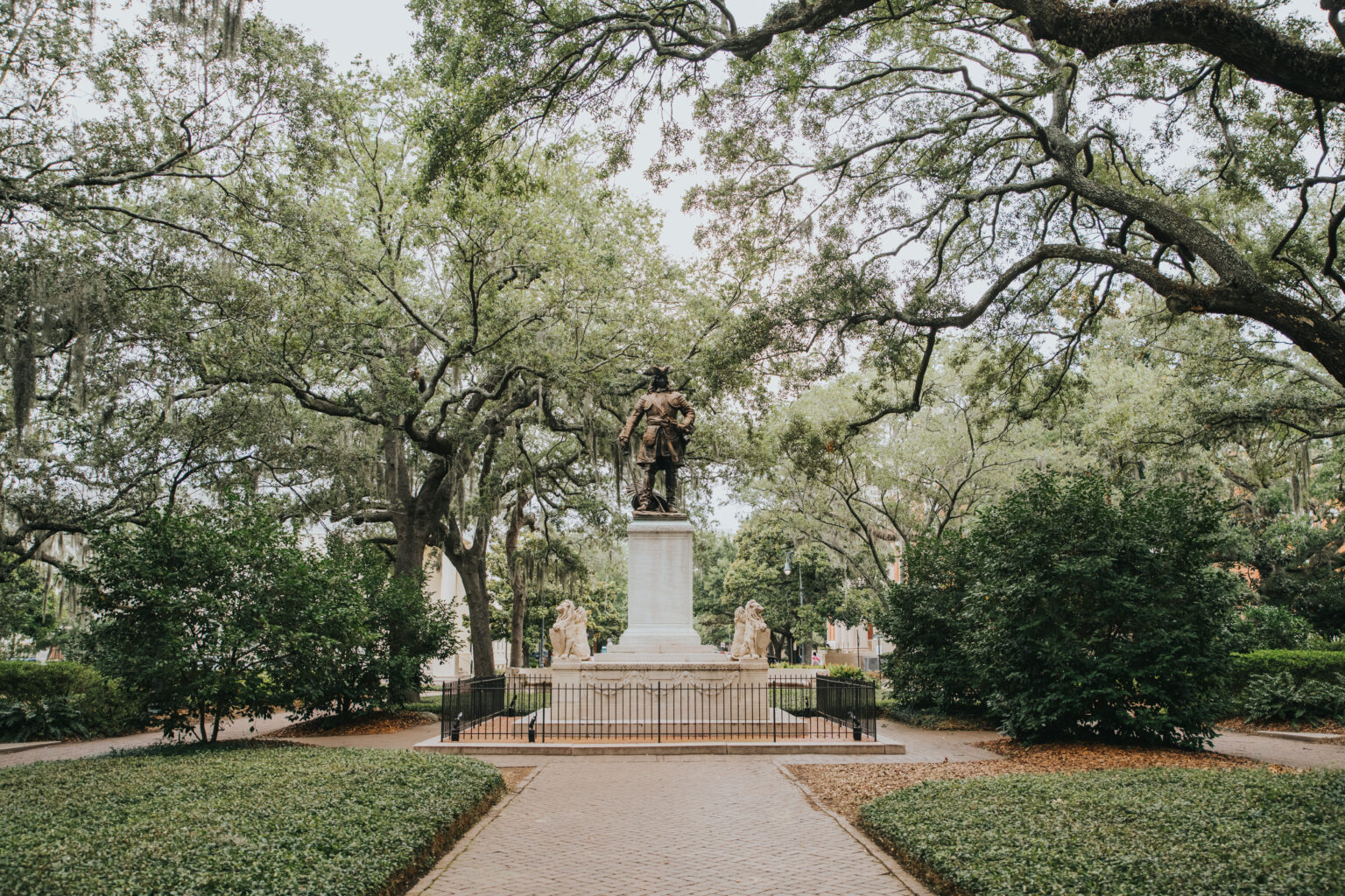 Elopement Wedding A statue stands on a tall pedestal at the center of a lush, green park with trees creating a canopy overhead. Perfect for intimate elopements, the statue is surrounded by a fence and set on a paved pathway. Four smaller statues are positioned symmetrically around the main statue, adding to the serene atmosphere. Elopements Inc