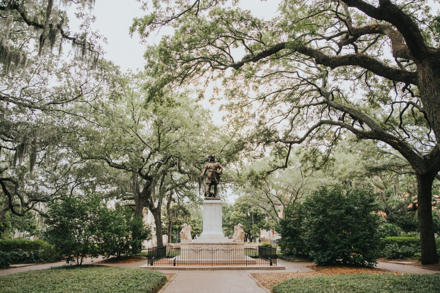 Elopement Wedding A bronze statue atop a stone pedestal stands surrounded by a black iron fence in a park filled with trees and lush greenery. The statue features a historical figure, and the trees have large, overarching branches creating a canopy effect—an ideal spot for elopements. Pathways and park benches are visible in the background. Elopements Inc