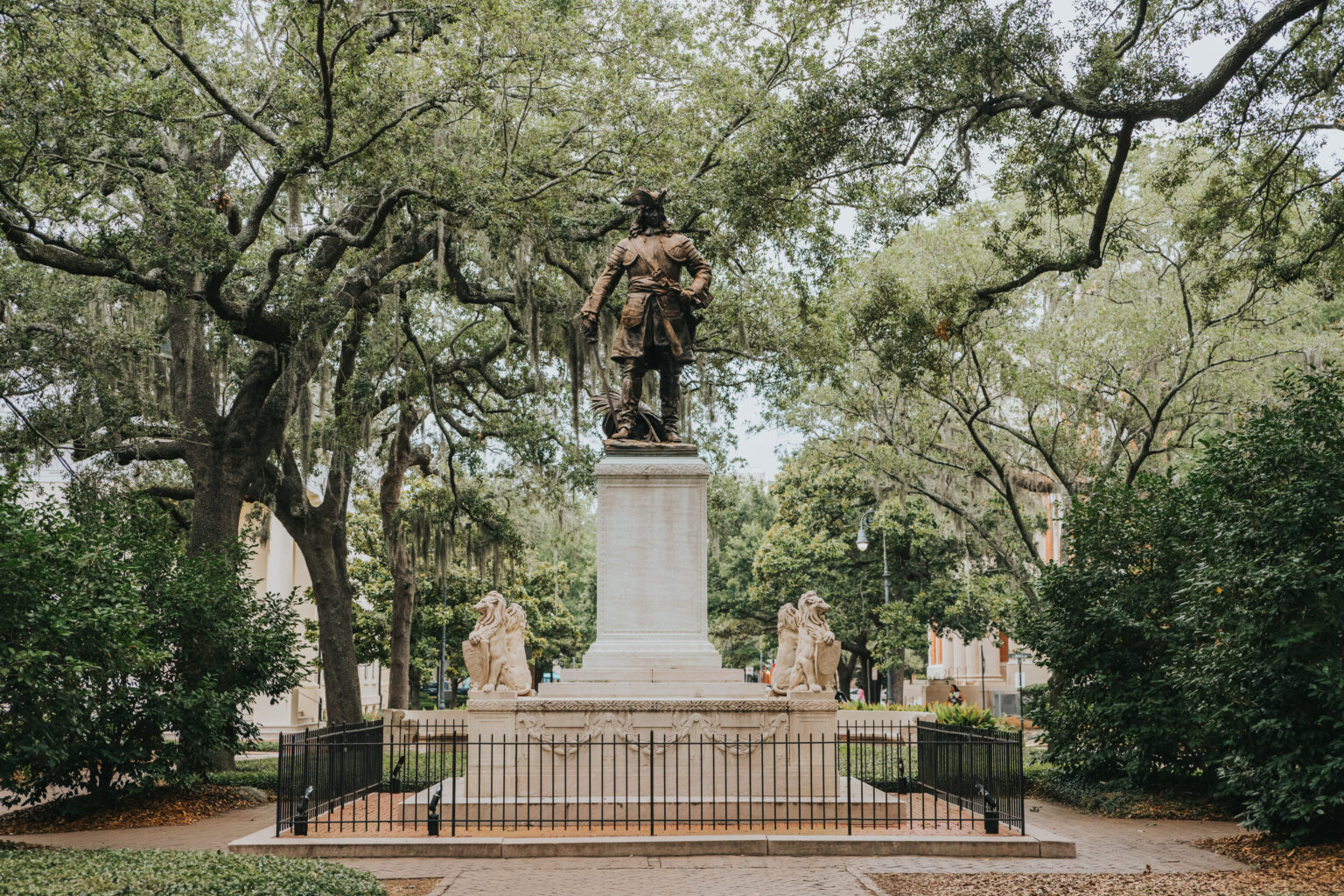 Elopement Wedding A bronze statue of a man standing on a tall pedestal, surrounded by four seated figures, is placed in the center of a fenced-off area. The monument is framed by large, sprawling trees and lush greenery, creating a serene park setting ideal for elopements with pathways leading to and surrounding the statue. Elopements Inc