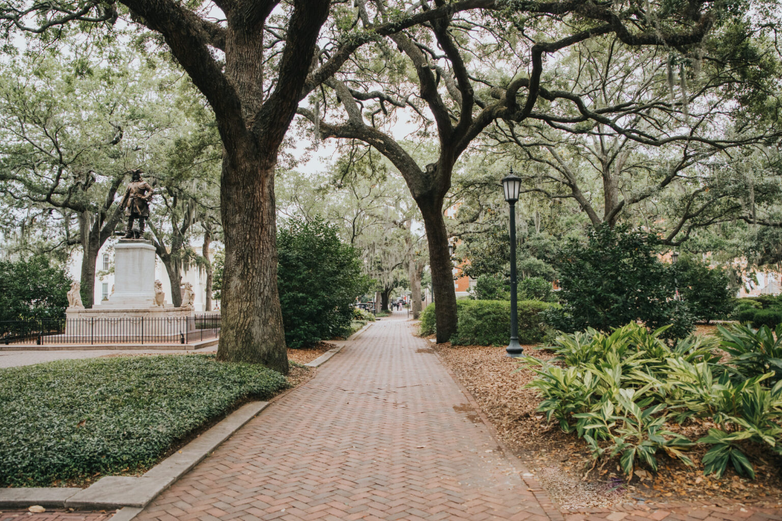 Elopement Wedding A serene brick pathway lined with lush greenery and large, arching trees leads to a distant statue on a white pedestal. The statue is in a small, fenced area perfect for an intimate elopement. A lamppost stands on the right side of the path. The atmosphere is calm and shaded by the dense canopy of leaves. Elopements Inc