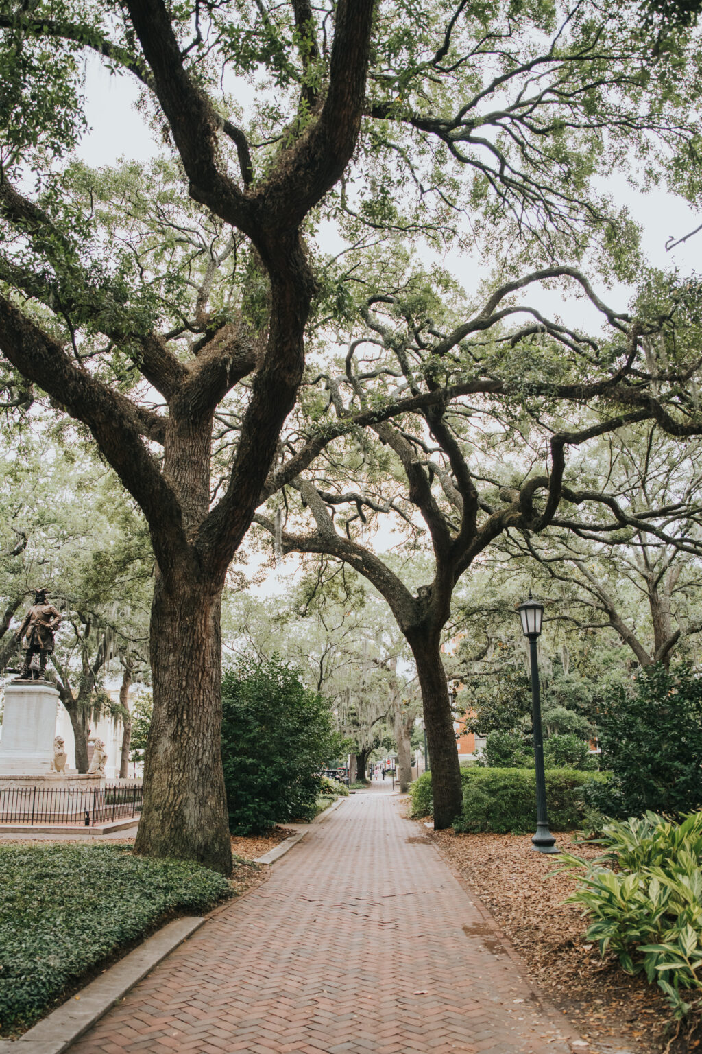 Elopement Wedding A tree-lined, brick-paved path in a park, with towering trees forming a canopy overhead. On the left, a statue stands behind a fence surrounded by greenery. A lamppost is visible on the right. Lush green bushes and patches of grass border the path, offering a serene backdrop perfect for an elopement. Elopements Inc