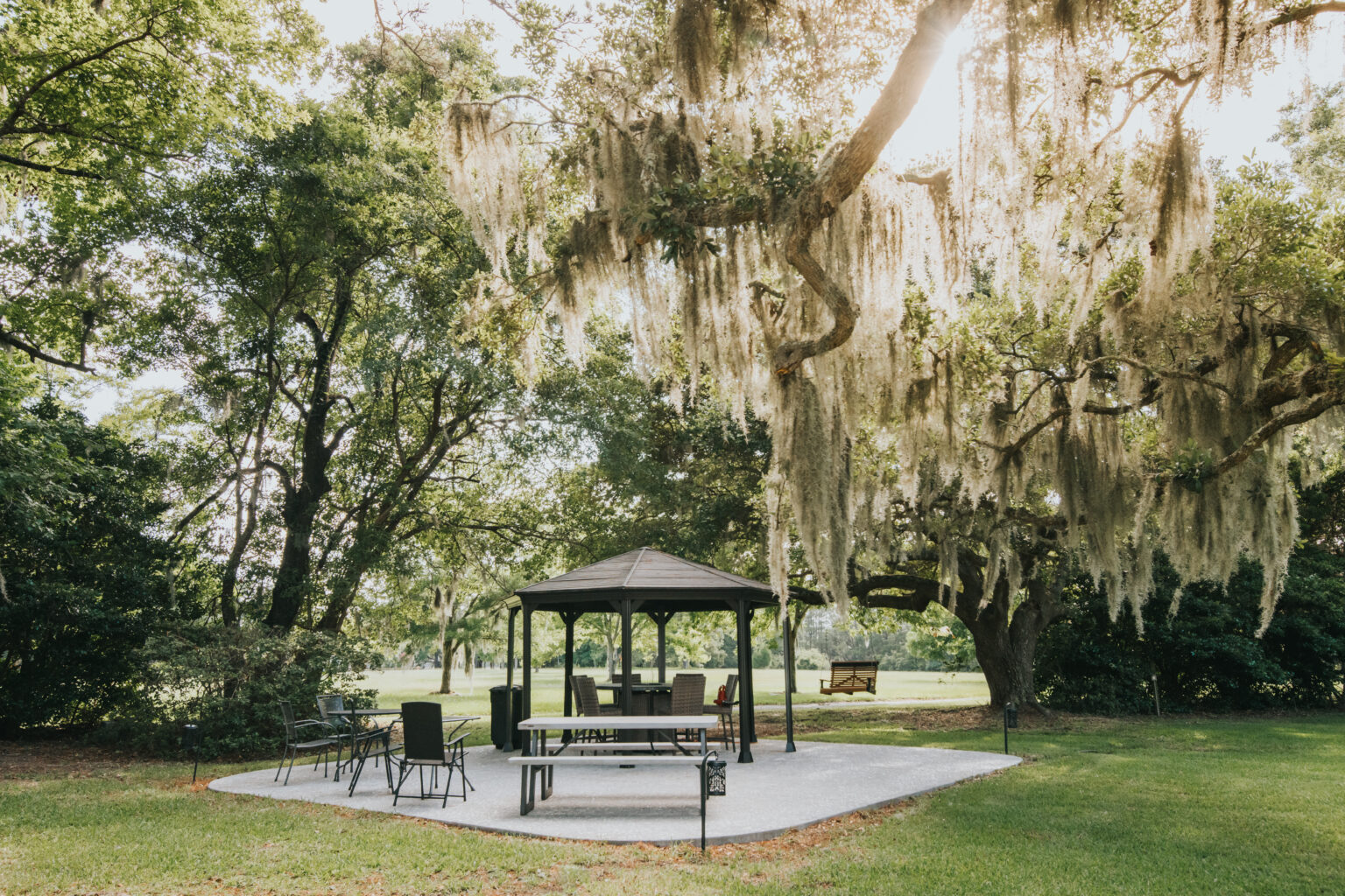 Elopement Wedding A serene park scene featuring a black gazebo placed on a concrete patio surrounded by lush, green grass and foliage. Spanish moss hangs from large, sprawling trees, and sunlight filters through the branches. This charming spot is perfect for intimate elopements with various seating options including a swing attached to a tree. Elopements Inc