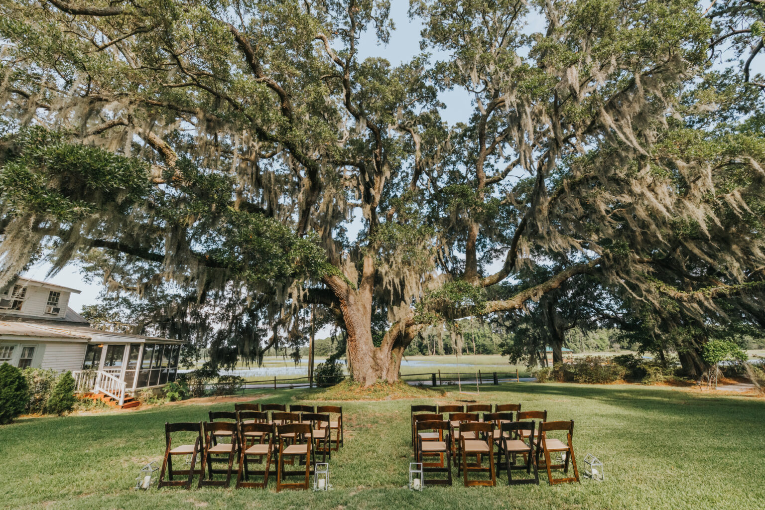 Elopement Wedding A large, scenic oak tree with sprawling branches draped in Spanish moss dominates a grassy outdoor setting. Rows of brown wooden chairs are arranged in two sections facing the tree, forming an aisle perfect for intimate elopements. In the background, a white house with a porch is visible on the left. Elopements Inc