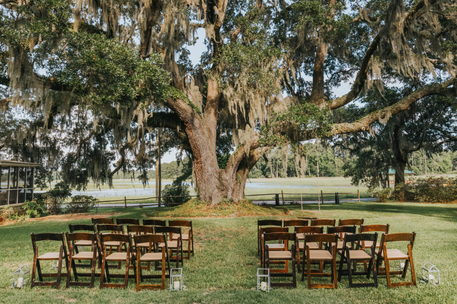 Elopement Wedding An outdoor wedding setup ideal for intimate elopements features rows of wooden chairs with beige cushions arranged on a grassy lawn. A large oak tree with hanging Spanish moss serves as the backdrop. Lanterns are placed along the aisle, and a scenic view of trees and a pond is visible in the background. Elopements Inc