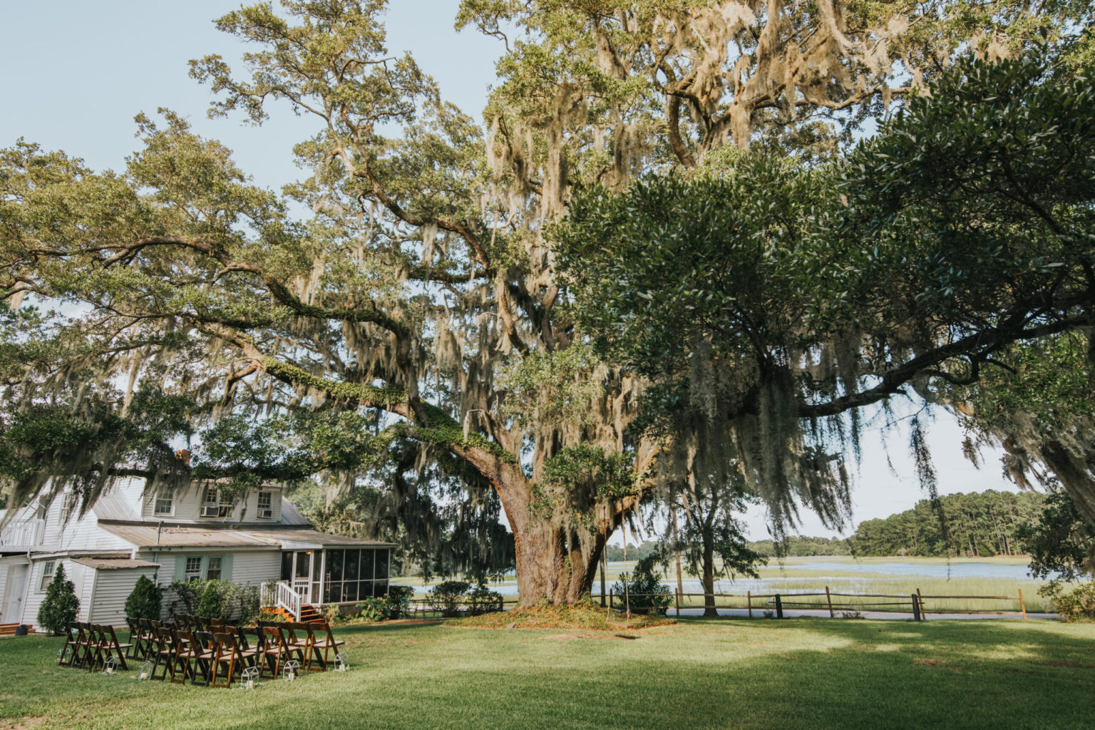 Elopement Wedding A large oak tree draped with Spanish moss looms over rows of brown wooden chairs set up on a well-manicured lawn, perfect for an elopement. A large white house with a screened porch and several windows is visible to the left. The background features a wooden fence and a serene body of water surrounded by greenery. Elopements Inc