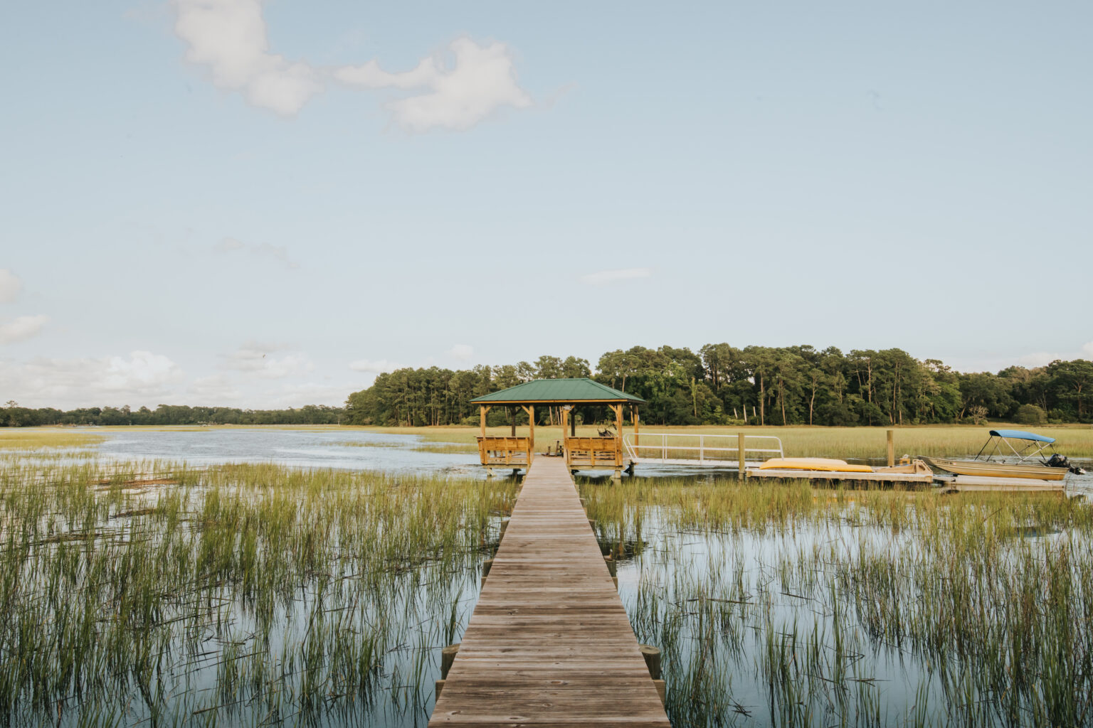 Elopement Wedding A wooden pier extends over calm water leading to a gazebo with a green roof, perfect for intimate elopements. Marsh grasses grow through the shallow water on both sides of the pier. In the background are dense green trees. A small dock to the right has a few boats moored. The sky is clear with a few white clouds. Elopements Inc