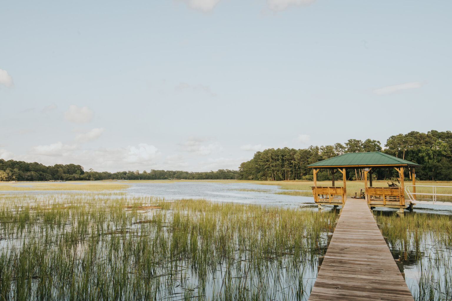 Elopement Wedding A wooden pier leads to a covered gazebo extending over a calm lake with marsh grasses. The lake is surrounded by green trees under a clear, light blue sky with a few scattered clouds. The scene is tranquil and nature-dominated, ideal for elopements or as a peaceful retreat and fishing spot. Elopements Inc