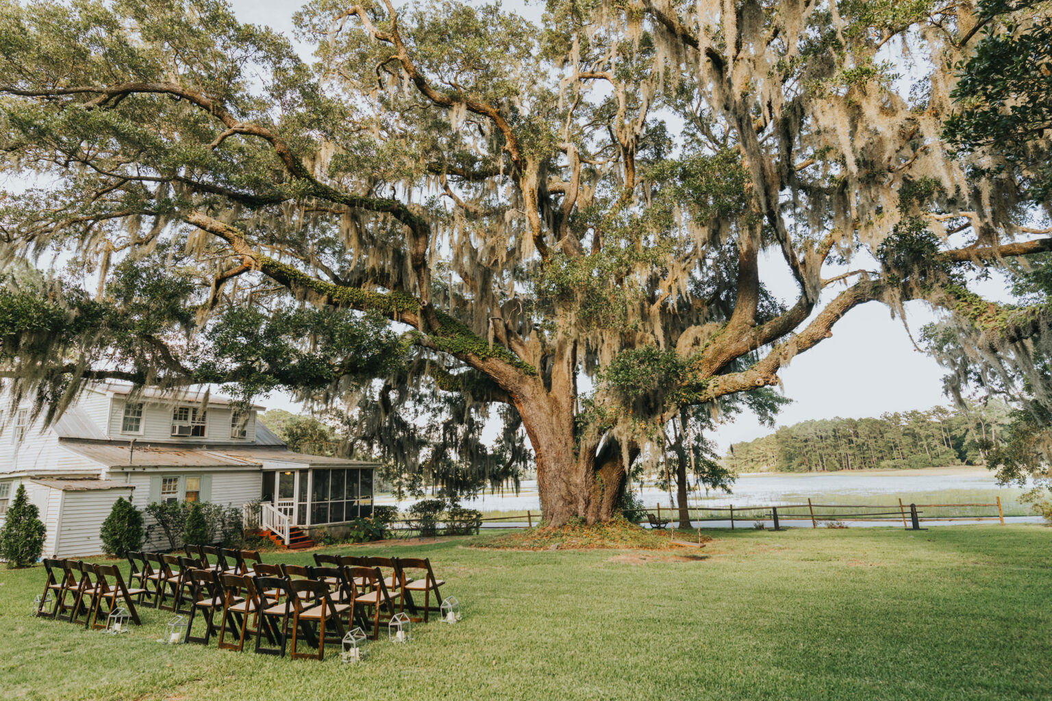 Elopement Wedding A large, picturesque tree with draping Spanish moss towers over an outdoor elopement setup. Wooden chairs are neatly arranged in rows on the lush grass, facing the tree. To the left, a small white house with a porch is visible. In the background, there's a body of water and a fence. Elopements Inc