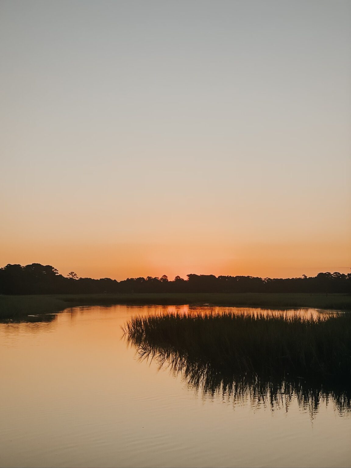 Elopement Wedding A serene sunset over a calm river with marsh grasses along the shore sets the perfect backdrop for an intimate elopement. The sky transitions from a soft orange near the horizon to a pale blue higher up, while the silhouettes of trees create a dark outline, gently reflecting on the water's surface. Elopements Inc