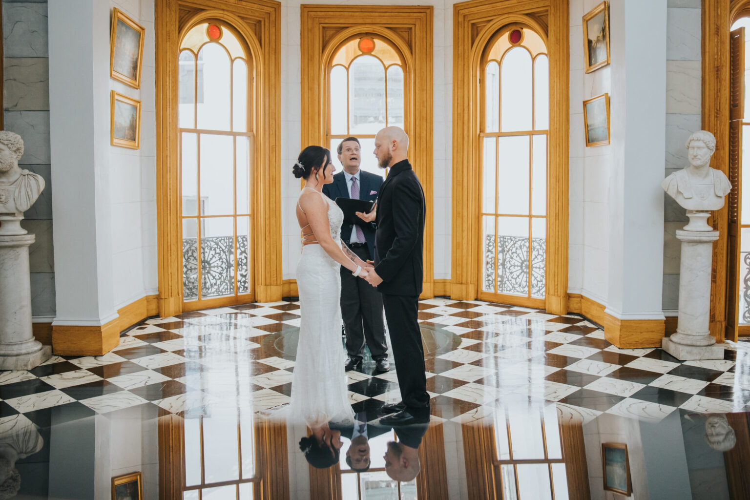 Elopement Wedding A couple stands facing each other holding hands, the bride in a white dress and the groom in a black suit, in a grand room with checkered floor, arched windows, and several paintings and busts on pedestals. An officiant stands behind them, conducting their intimate elopement ceremony. Elopements Inc