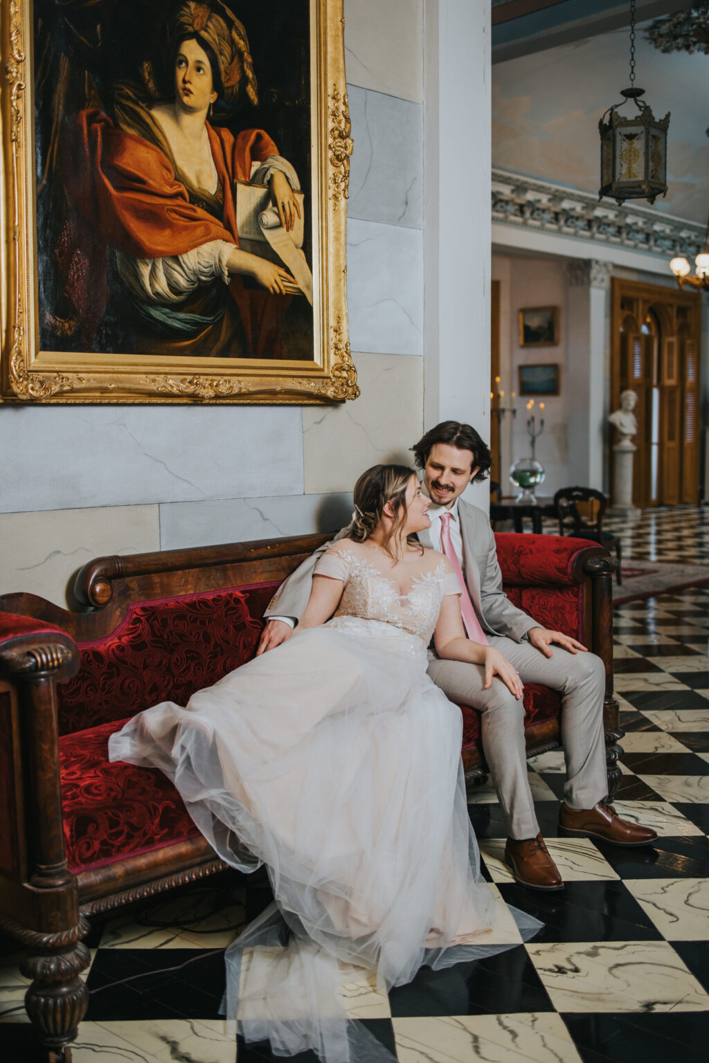 Elopement Wedding A bride and groom, having just eloped, sit on a red velvet couch in an elegant room. The bride, in a white lace gown, leans on the groom, who wears a beige suit. A large painting of a person in a red garment hangs above them. The room features black-and-white checkered flooring and ornate decorations. Elopements Inc