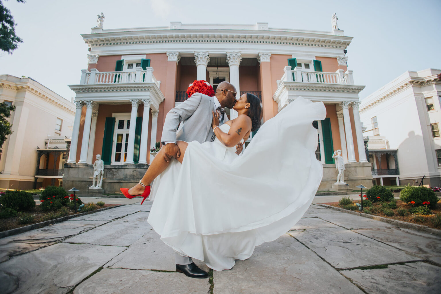 Elopement Wedding A groom in a gray suit lifts and kisses his bride, who wears a flowing white dress, red shoes, and a veil, in front of a grand building with columns, green shutters, and statues. The couple's elopement adds an air of spontaneity to the scene as the bride's dress billows gracefully. Elopements Inc