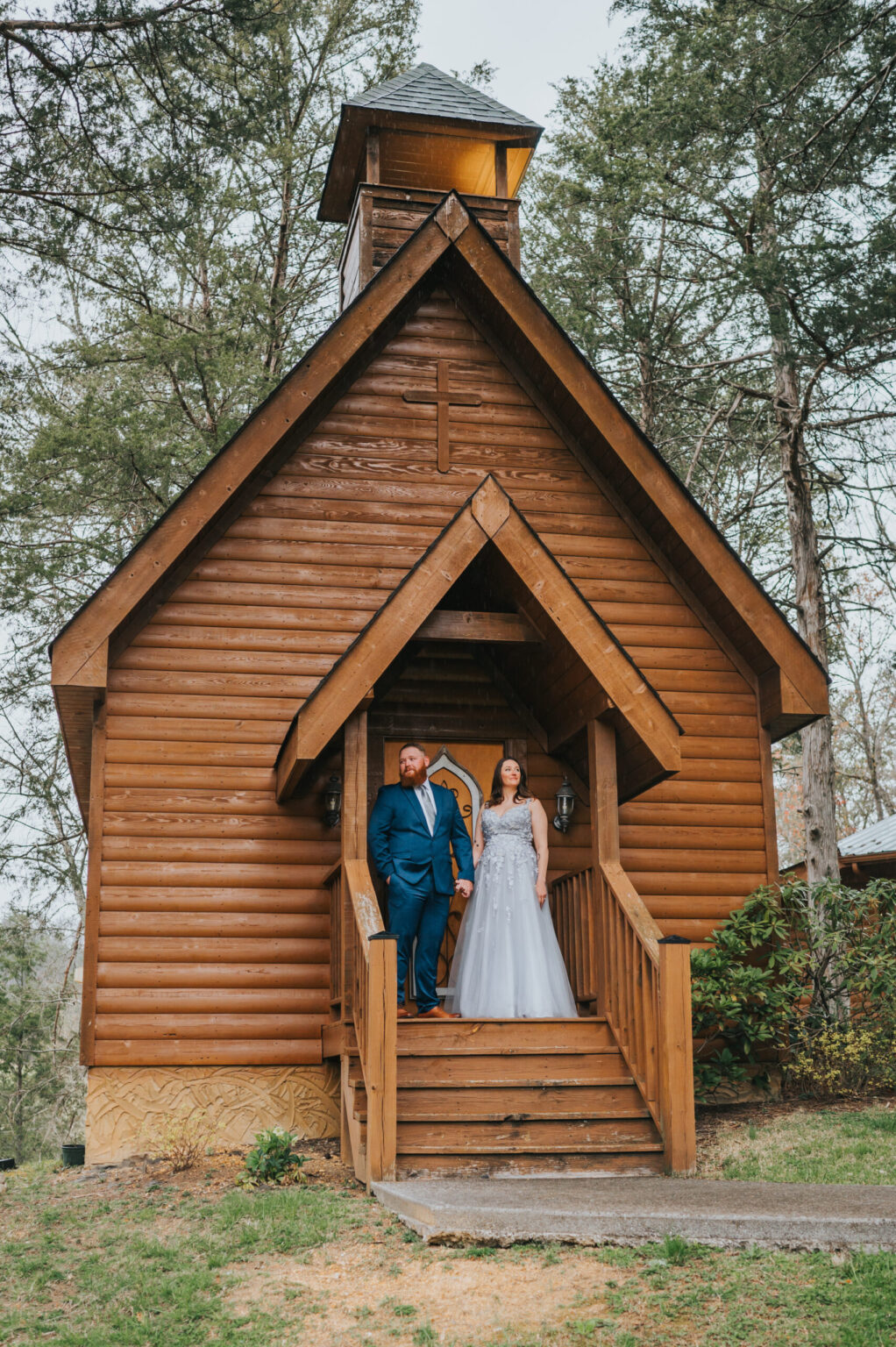 Elopement Wedding A couple stands on the steps of a charming wooden chapel with a sloped roof, surrounded by tall trees. The groom is in a blue suit and the bride in a white gown. Both gaze into the distance, exuding a serene and joyful demeanor after their intimate elopement. The setting is picturesque and natural. Elopements Inc