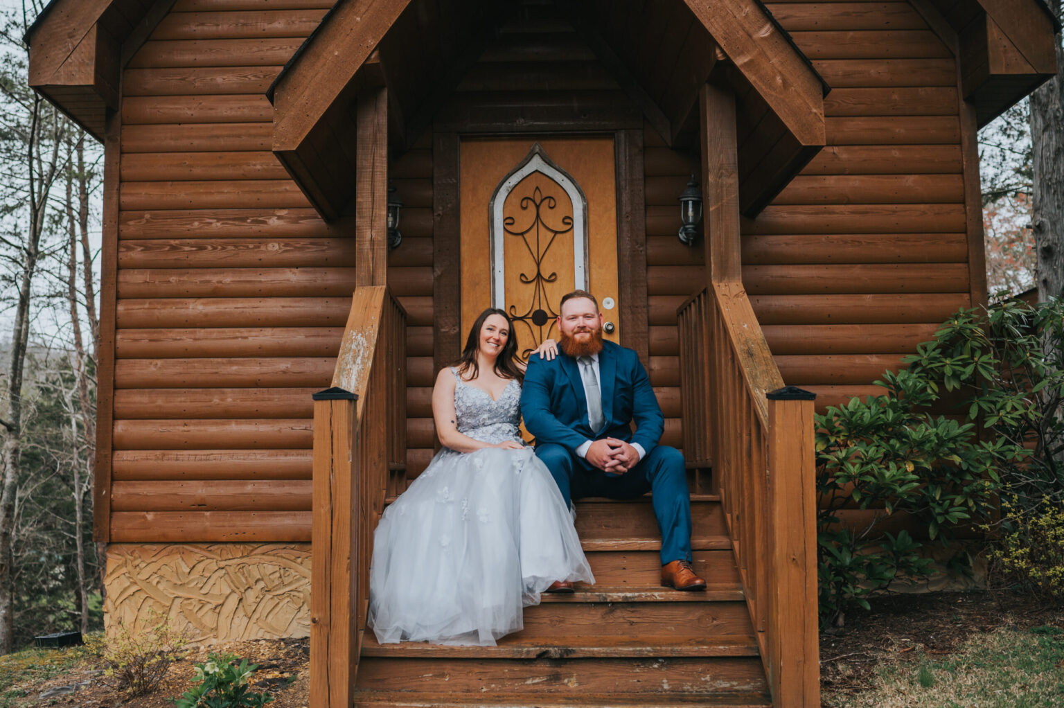 Elopement Wedding A smiling couple, recently eloped, sits on the wooden steps of a rustic cabin. The woman wears a light grey sleeveless gown, while the man is dressed in a blue suit. The cabin door features ornate ironwork. Green foliage is visible to the right, and tree trunks can be seen in the background. Elopements Inc