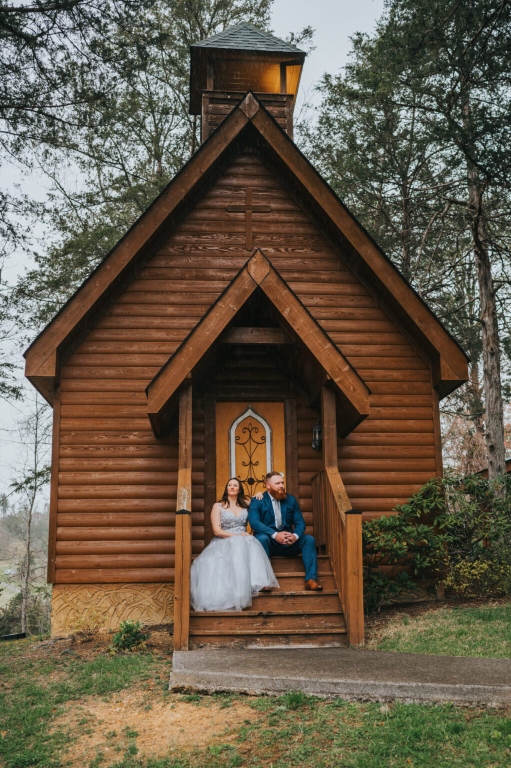 Elopement Wedding A couple dressed in wedding attire sits on the wooden steps of a small, rustic chapel surrounded by trees. The bride wears a white gown, and the groom dons a blue suit. The chapel has a tall, pointed roof and a wooden door with a decorative glass panel. The serene backdrop is perfect for their elopement. Elopements Inc