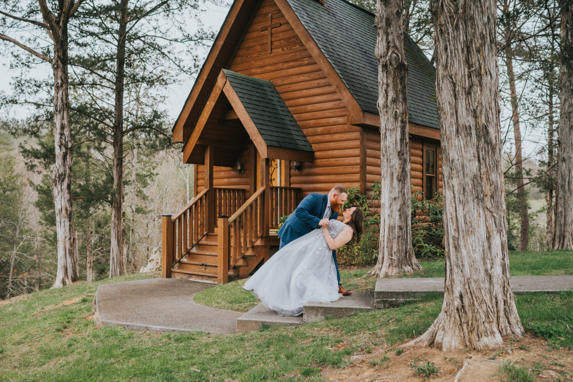 Elopement Wedding The groom in a blue suit and the bride in a white gown share a kiss as he dips her in front of a small, charming wooden chapel. Surrounded by trees and greenery, the rustic setting adds to the romantic backdrop, capturing their tender elopement moment in a serene outdoor setting. Elopements Inc