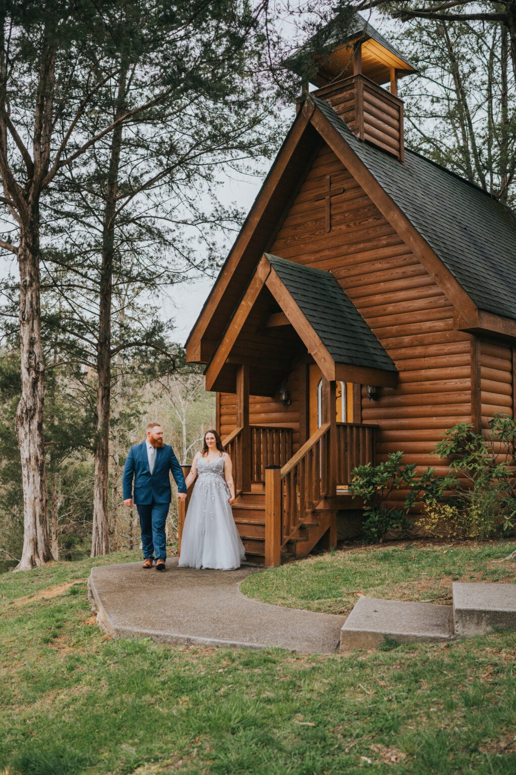 Elopement Wedding A couple stands in front of a small wooden chapel surrounded by tall trees. The man, dressed in a blue suit, holds hands with the woman in a white wedding gown. The chapel has a pitched roof, a small steeple, and a porch. Ideal for elopements, a winding concrete path leads up to the entrance. Elopements Inc