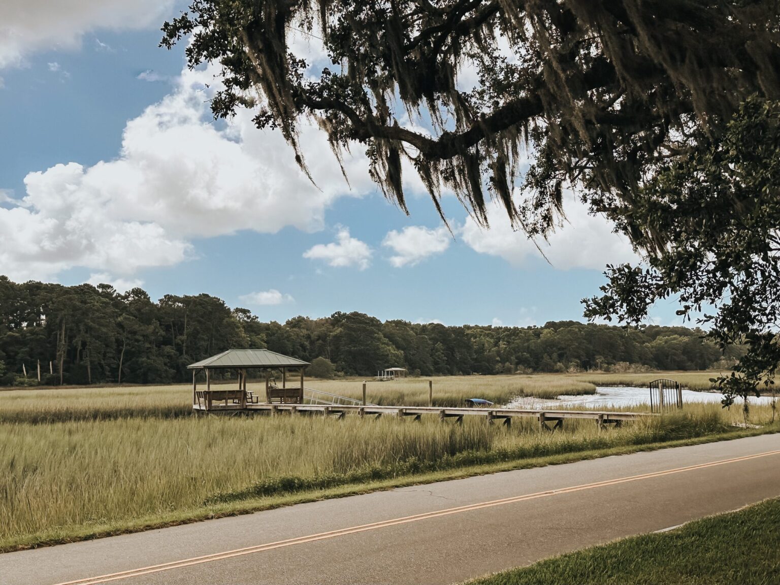 Elopement Wedding A tranquil scene features a small wooden gazebo over water on a dock, perfect for an intimate elopement. The dock extends from the lush, green marshland. A two-lane road runs along the foreground with trees draped in Spanish moss hanging from the top, under a partly cloudy sky. Forested land fills the background. Elopements Inc