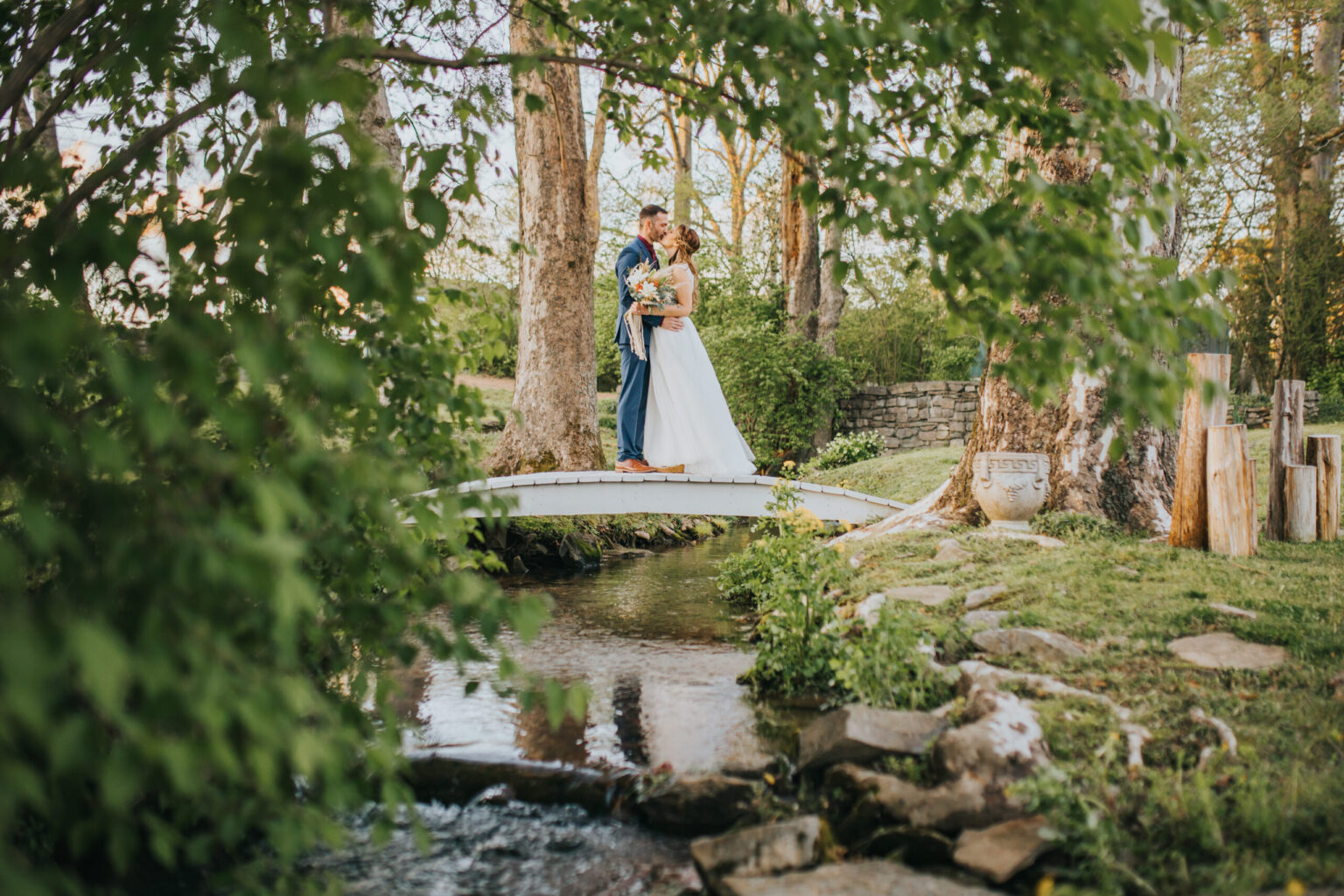 Elopement Wedding A bride and groom, who chose to elope, stand on a small white bridge over a serene creek in a lush, green garden. They are embracing, and the bride holds a bouquet of flowers. Trees and greenery surround them, creating a peaceful, romantic atmosphere. Stone and wooden elements accent the natural setting. Elopements Inc