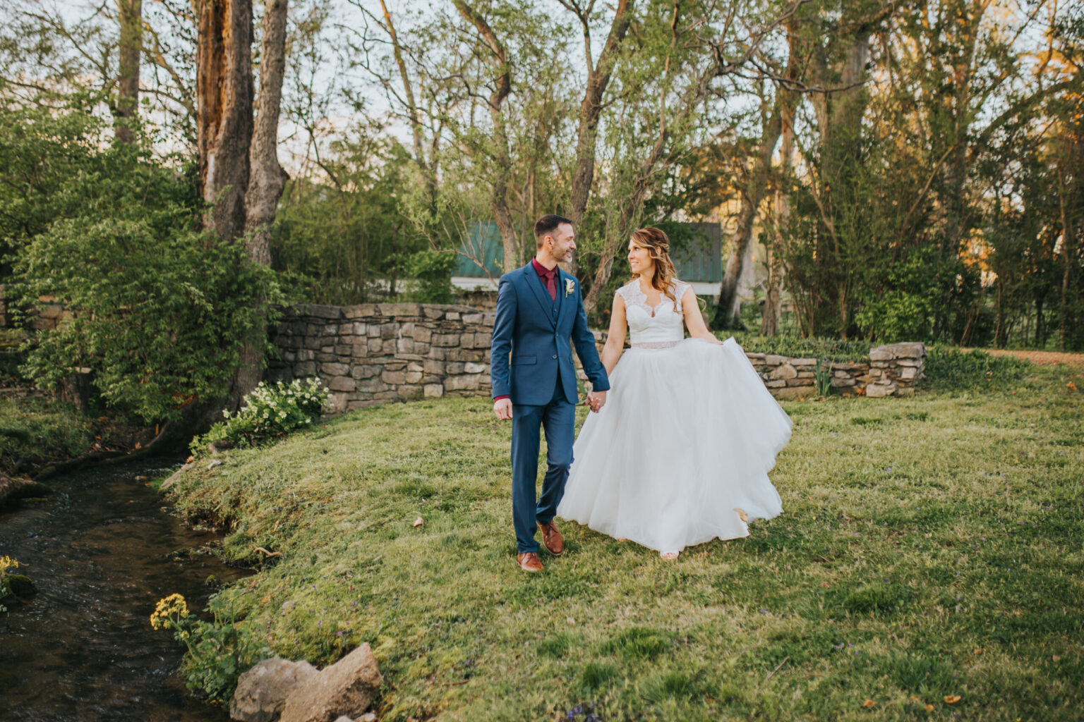 Elopement Wedding A couple walks hand-in-hand on a grassy area near a small creek, having just eloped. The woman wears a white bridal gown, and the man sports a blue suit with a dark red tie. The background features a low stone wall, lush green trees, and a clear sky, suggesting a serene outdoor setting, possibly a garden or park. Elopements Inc