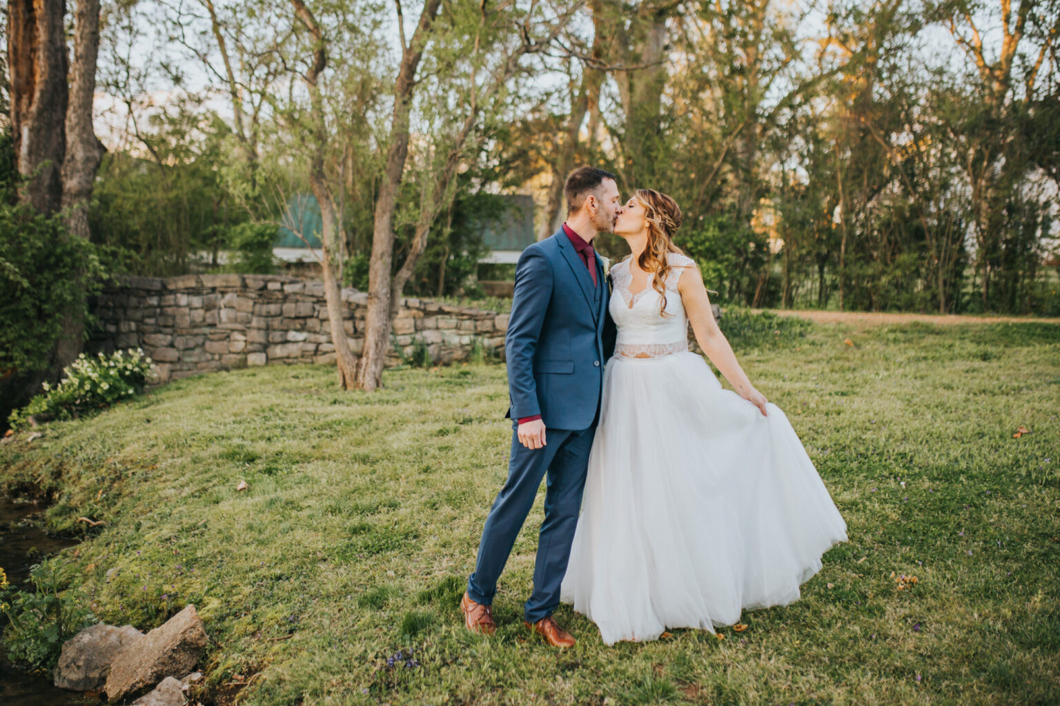 Elopement Wedding A bride in a white gown and a groom in a blue suit share a kiss outdoors in a grassy area with a stone wall and trees in the background. The bride holds up part of her dress, and sunlight filters through the trees, casting a warm glow on this intimate elopement. Elopements Inc