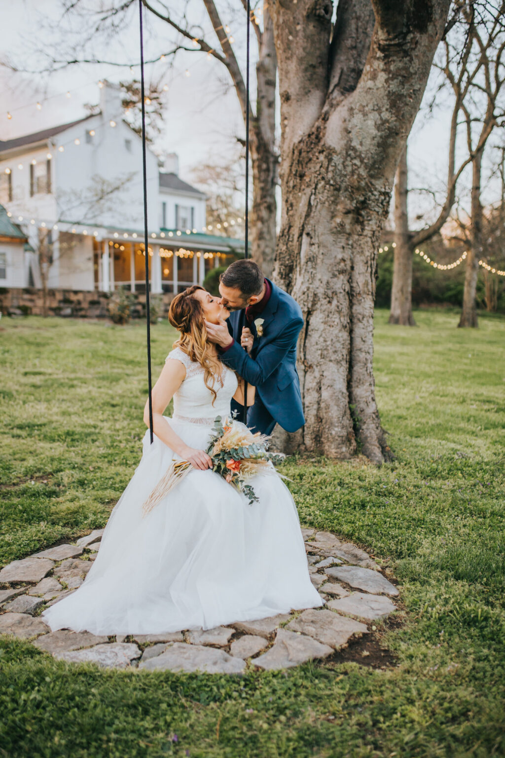 Elopement Wedding A bride in a white wedding dress holds a bouquet while sitting on a swing. Her groom, in a dark suit, leans down to kiss her. They are outdoors, with a stone circle around the swing base. In the background, there is a large tree, house, and string lights creating the perfect ambiance for their intimate elopement. Elopements Inc