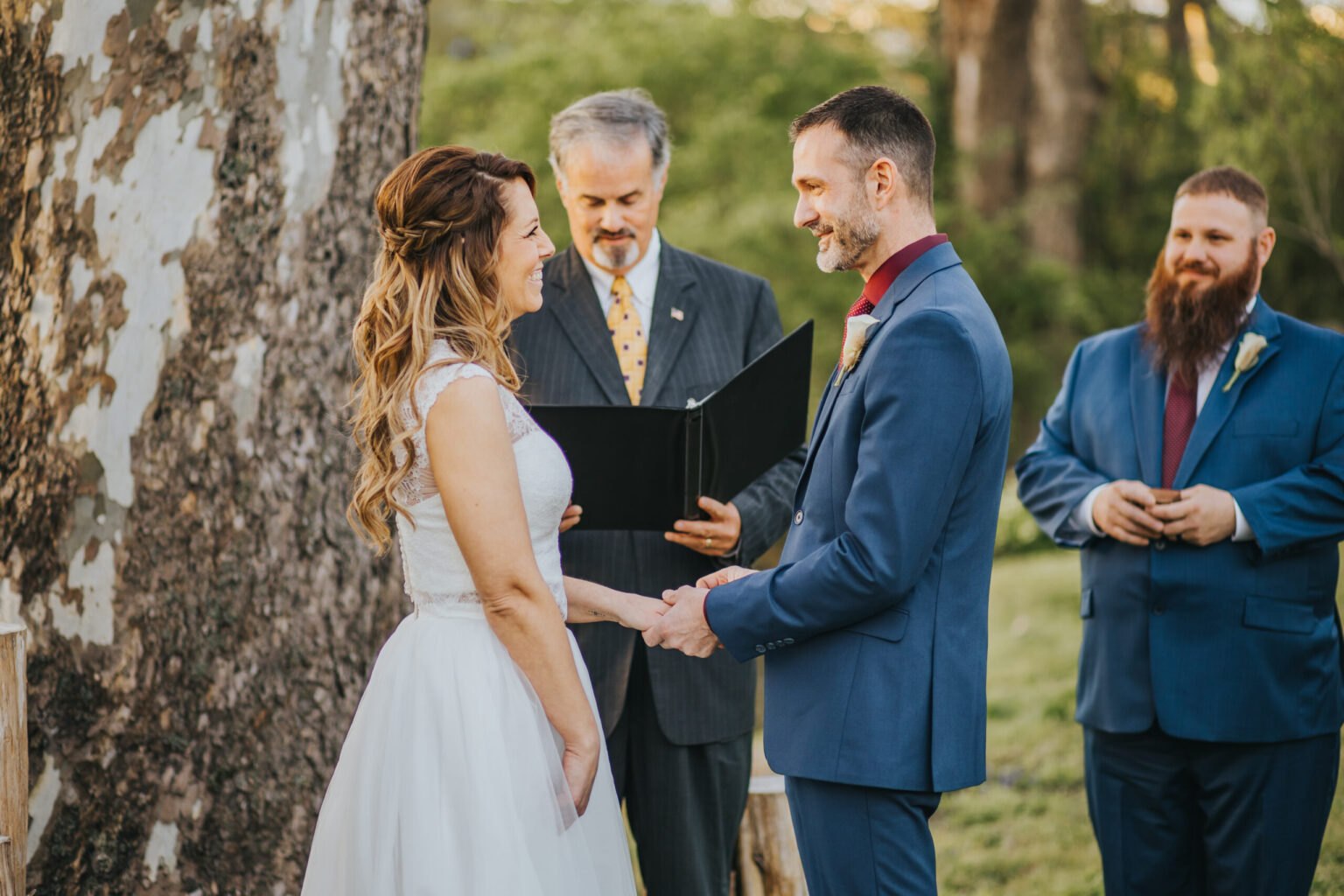 Elopement Wedding A bride and groom exchange vows during their elopement outdoors under a large tree. The bride wears a white dress, the groom a blue suit with a red tie. A man officiates the ceremony, holding a black folder, while another man in a blue suit with a long beard stands to the right, hands clasped together. Elopements Inc