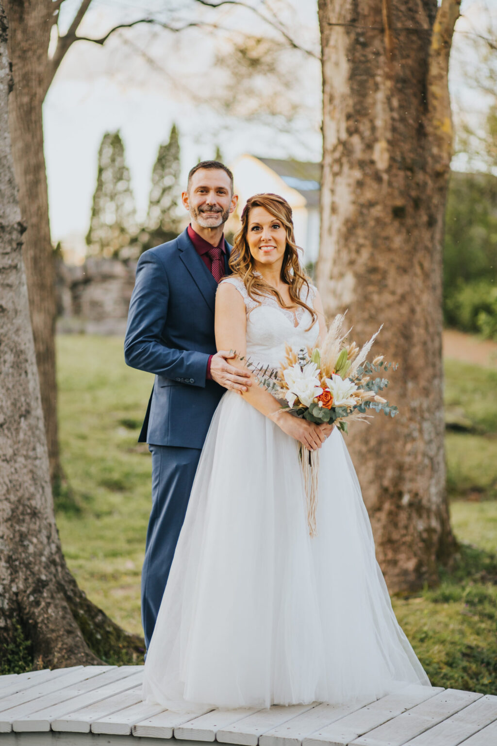 Elopement Wedding A bride and groom stand outdoors on a small wooden platform between two large trees. The groom wears a blue suit and burgundy shirt, while the bride, who is holding a bouquet of mixed flowers, wears a white gown. Smiling brightly during their intimate elopement, the bride stands slightly in front of the groom. Elopements Inc