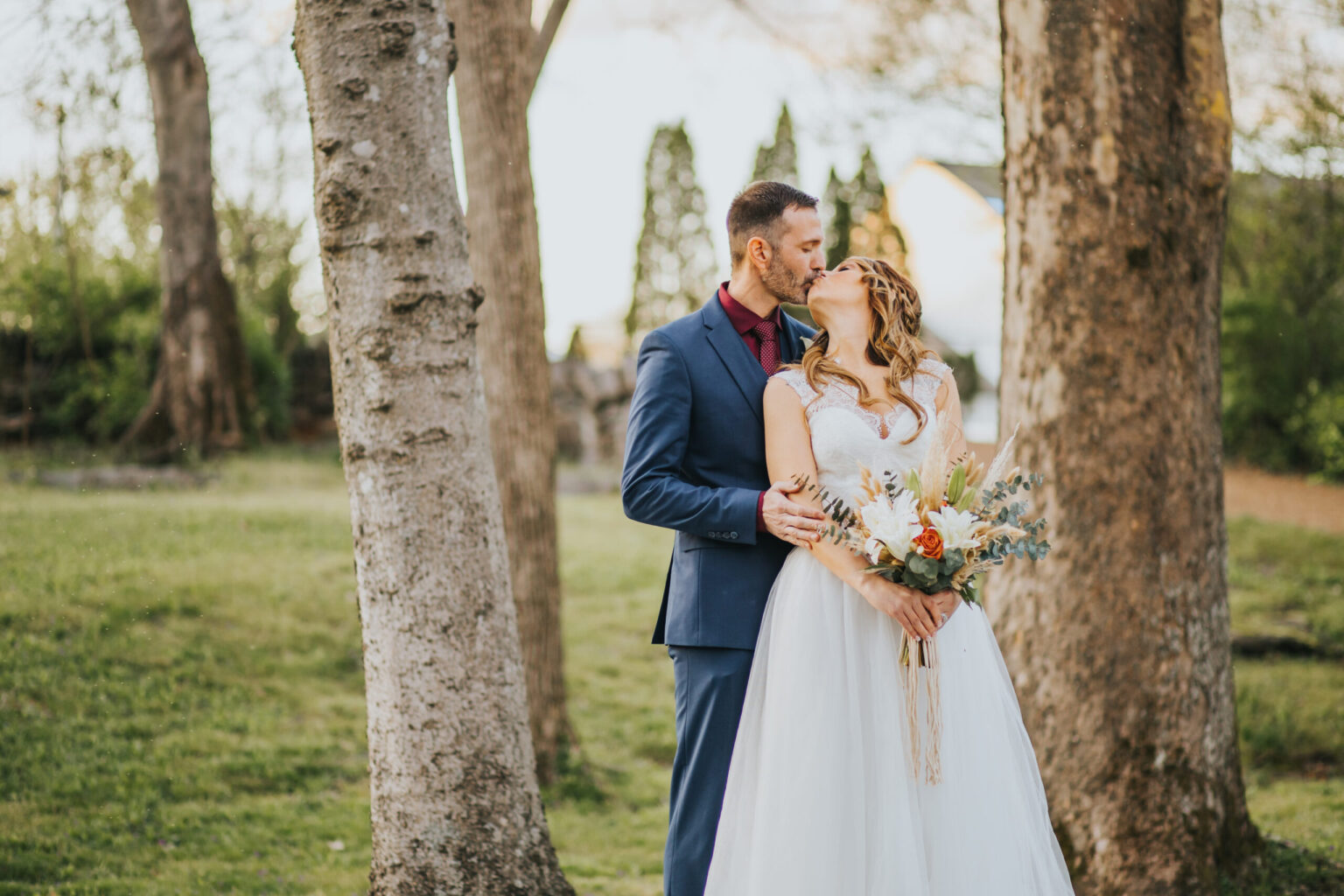 Elopement Wedding A groom in a blue suit and a bride in a white gown share a kiss while standing among trees in a lush, green outdoor elopement setting. The bride is holding a bouquet of flowers and looking up at the groom, who gently embraces her from behind. Sunlight filters through the trees, adding magic to their secret moment. Elopements Inc