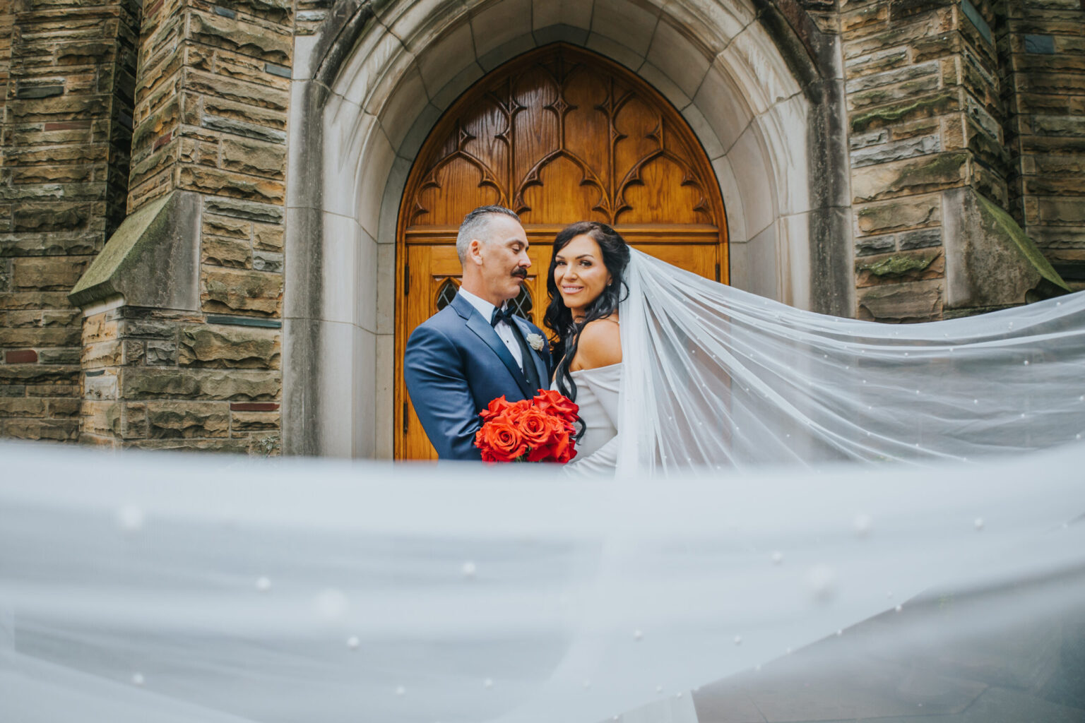 Elopement Wedding A bride and groom who chose to elope stand in front of a wooden church door with stone walls. The groom, wearing a blue suit and bowtie, holds a bouquet of red roses. The bride, in an off-the-shoulder white dress with a long, flowing veil, smiles while looking over her shoulder at the camera. Elopements Inc