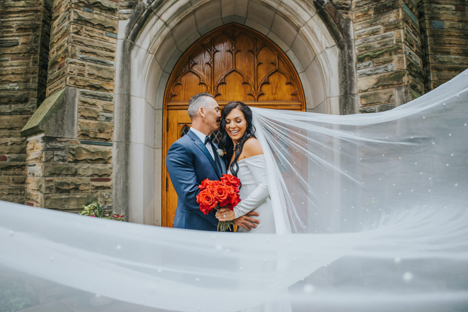 Elopement Wedding A bride and groom stand in front of a stone church entrance with arched wooden doors. The groom, in a blue suit, embraces the bride, who is in an off-shoulder white gown with a long, flowing veil. She holds a bouquet of red roses as both look joyful. Their elopement moment is captured as her veil billows elegantly around them. Elopements Inc