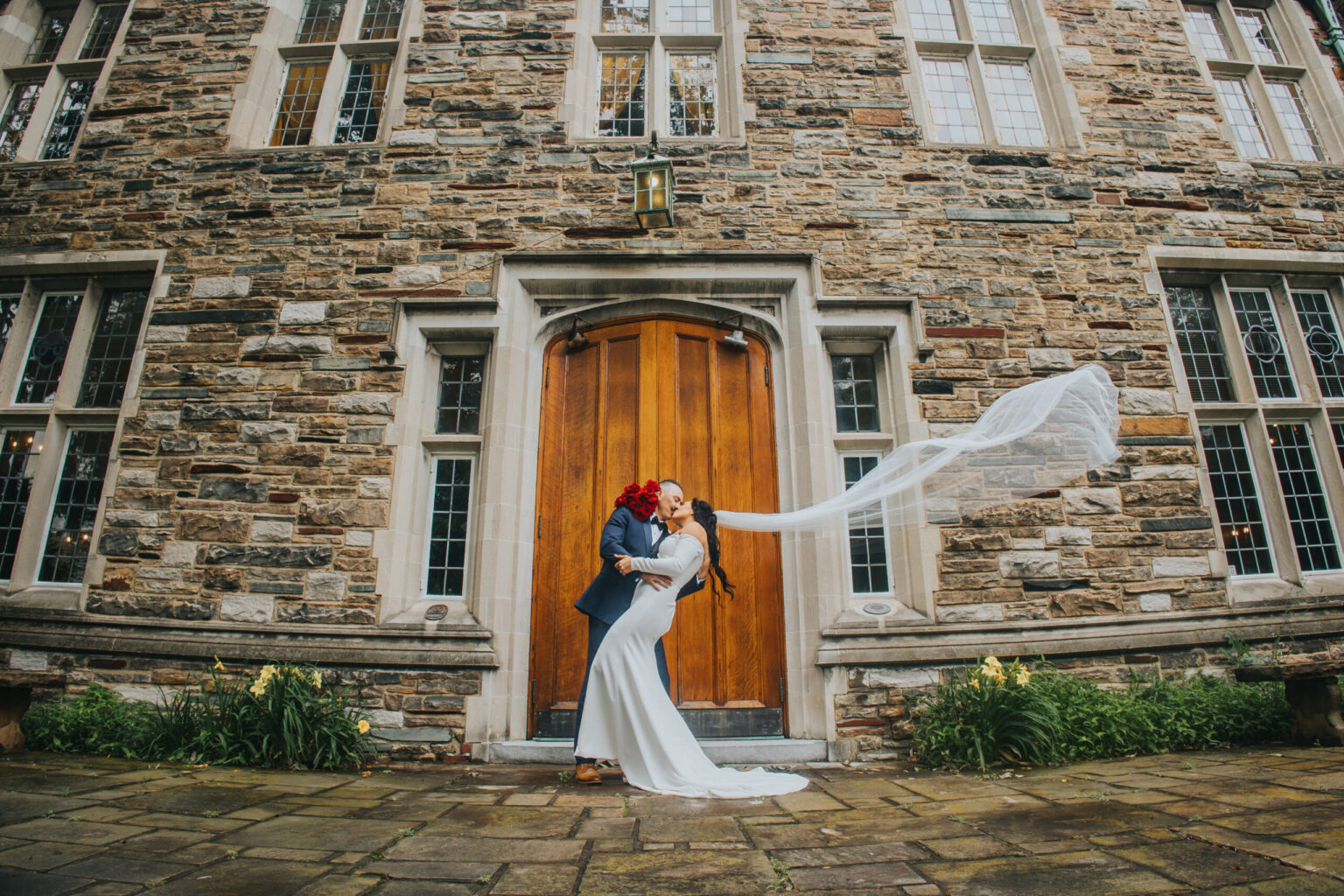 Elopement Wedding A newlywed couple embraces and kisses in front of a large wooden door on a stone building, having just eloped. The bride's veil is flowing in the wind, creating a dramatic effect. The groom wears a suit, and the bride is in a white dress. The ground is paved with stones, and there are plants flanking the entrance. Elopements Inc