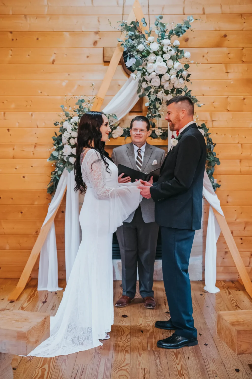 Elopement Wedding A couple is getting married in a wooden chapel. The bride, dressed in a long-sleeved white lace gown, and the groom, wearing a black suit with a red bow tie, stand facing each other holding hands. A man in a suit officiates the ceremony. They are positioned under a triangular floral arch adorned with white roses. Elopements Inc