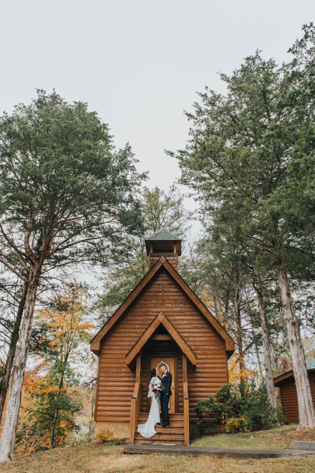 Elopement Wedding A bride and groom stand in front of a small wooden chapel surrounded by tall trees. The bride is wearing a white dress and holding a bouquet, while the groom is in a dark suit. The chapel, with a steep roof and small bell tower, is set in a serene, wooded area with autumn foliage. Elopements Inc