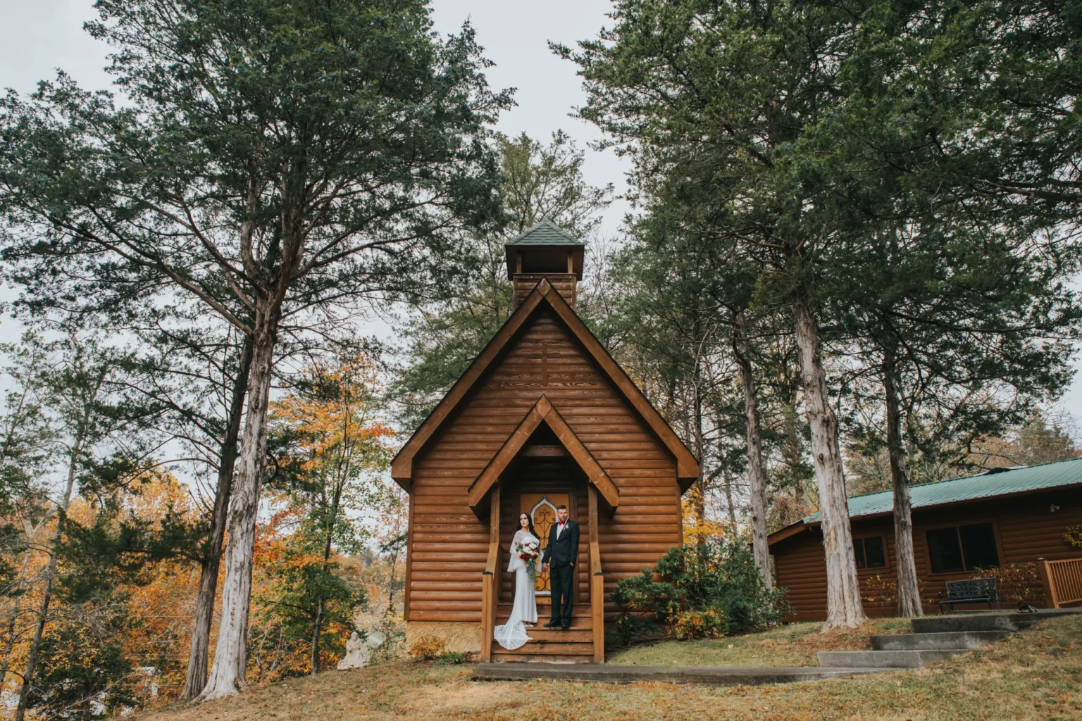 Elopement Wedding A bride in a white dress and a groom in a dark suit stand on the steps of a small wooden chapel surrounded by trees with autumn foliage. The chapel has a steeple and a cross at the top. A cabin is visible in the background to the right. The couple is smiling and holding hands. Elopements Inc