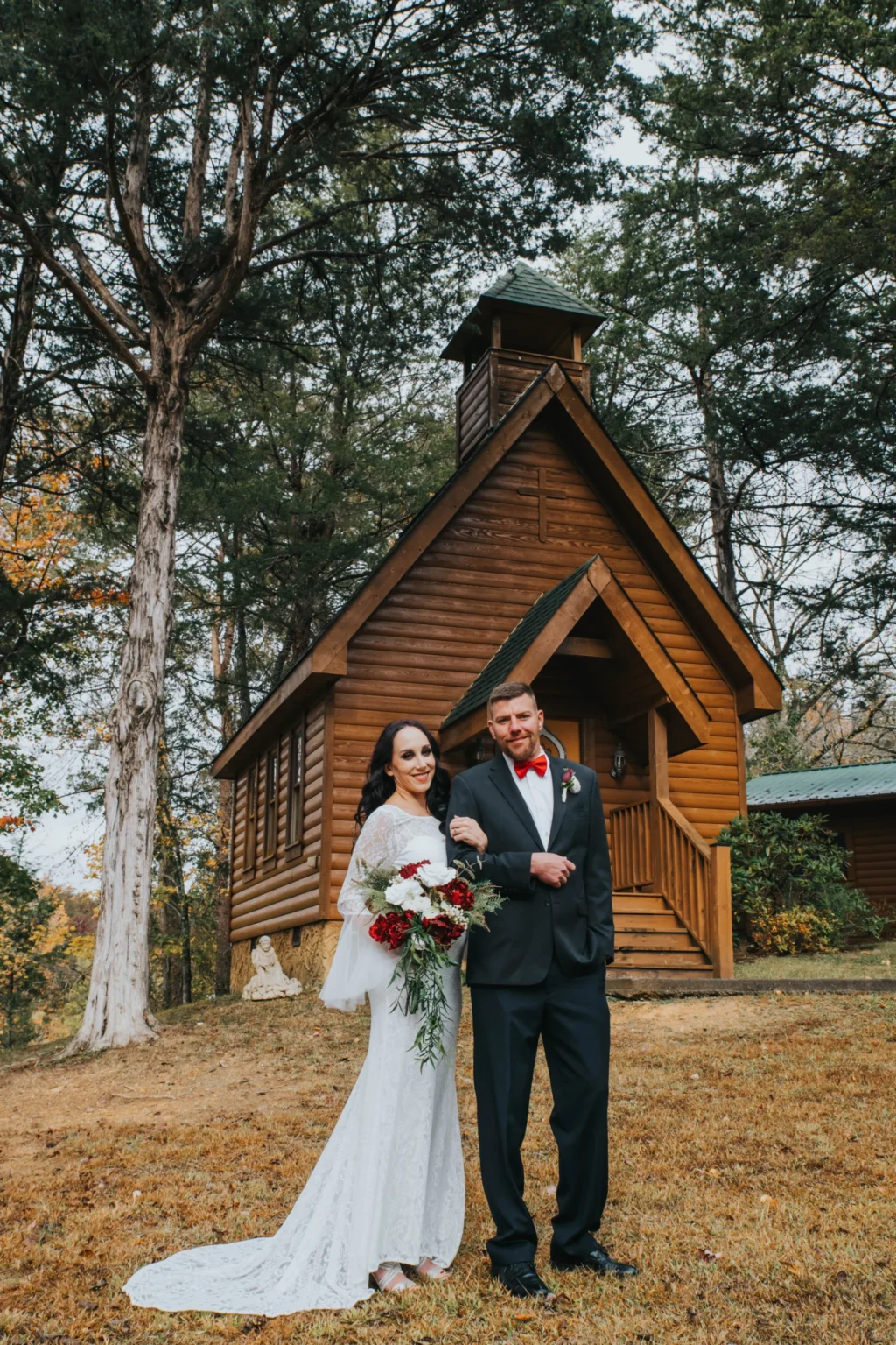 Elopement Wedding A bride in a white wedding dress holding red and white flowers and a groom in a black suit with a red bow tie stand arm in arm outside a rustic wooden chapel surrounded by tall trees. The groom smiles while the bride, also smiling, looks ahead. It is a serene, natural setting. Elopements Inc