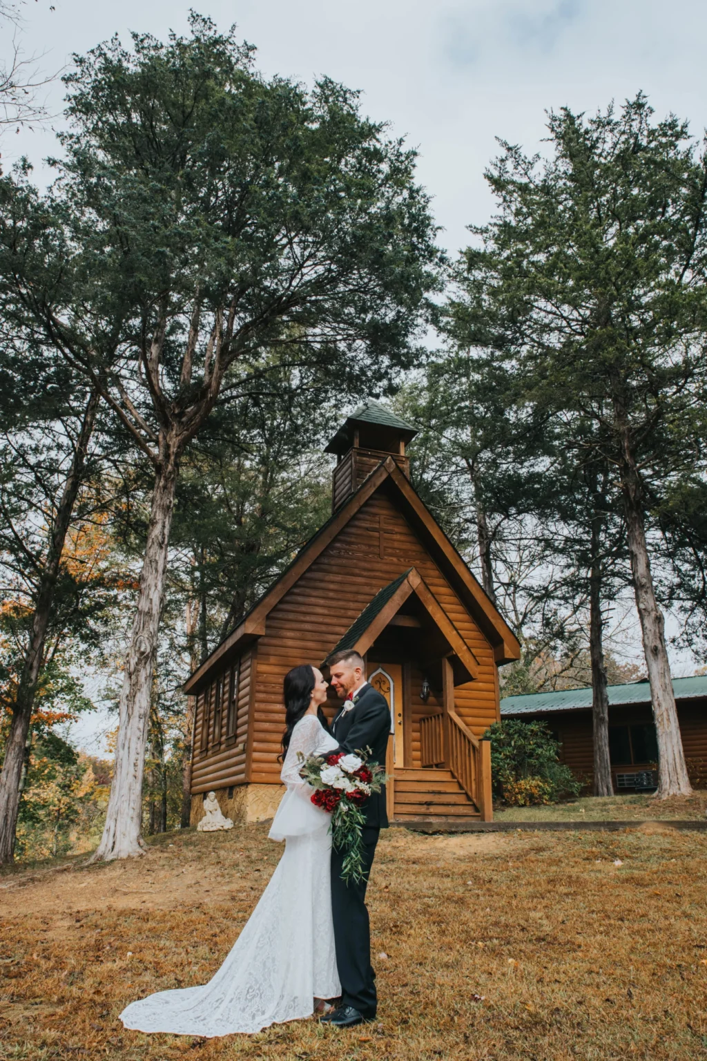 Elopement Wedding A bride in a long white lace dress and a groom in a black suit stand closely together, holding a bouquet of red and white flowers. They are in front of a small wooden chapel with a steeple, surrounded by tall trees and greenery, on a slightly overcast day. Elopements Inc