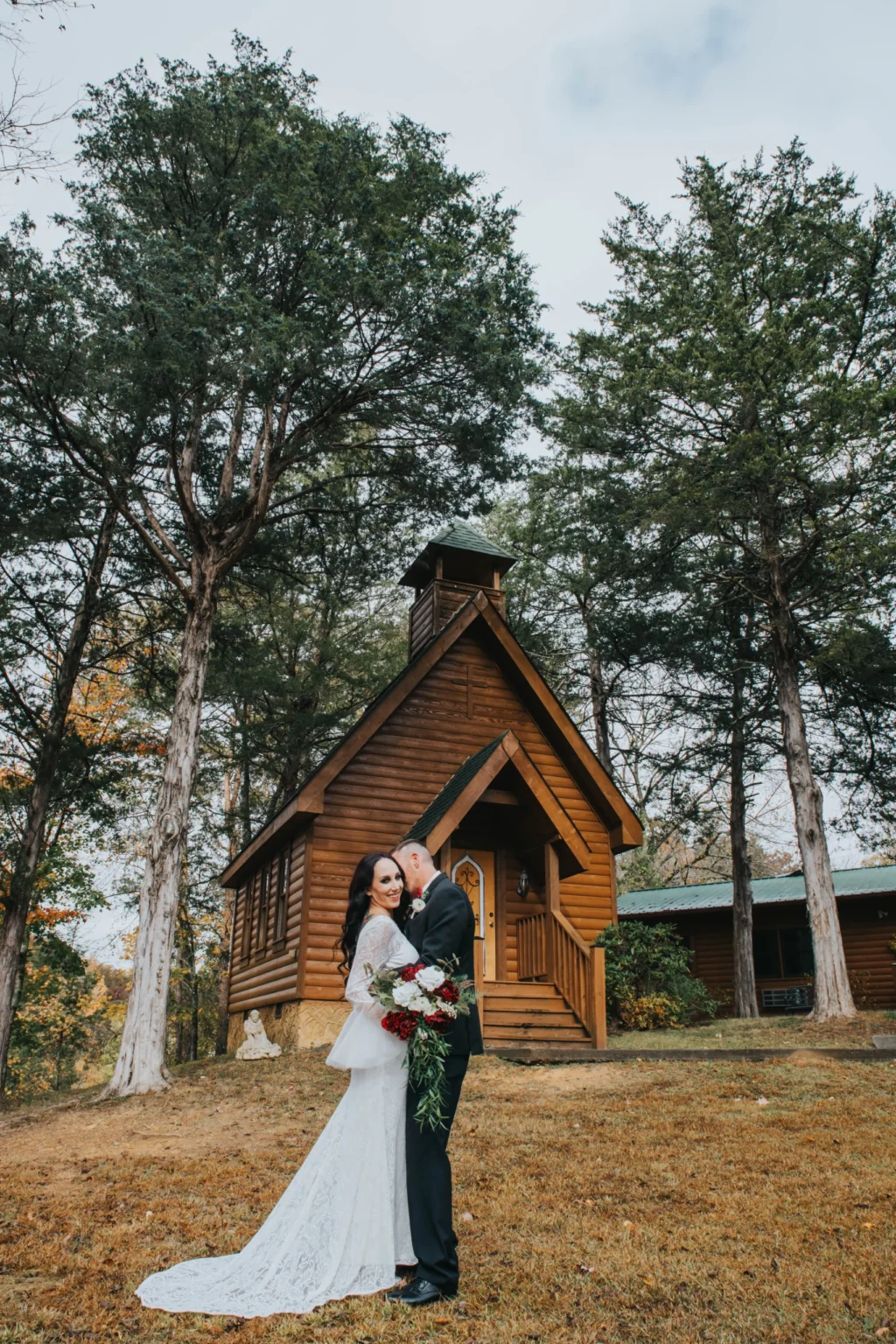 Elopement Wedding A bride and groom stand embracing in front of a small, rustic wooden chapel surrounded by tall trees. The bride wears a long, white lace wedding gown and holds a bouquet of red and white flowers. The groom is dressed in a black suit and kisses the bride's forehead. The scene is serene and picturesque. Elopements Inc