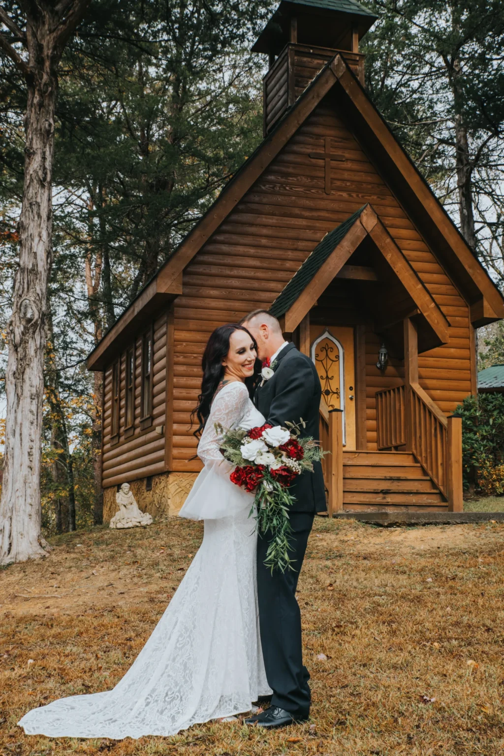 Elopement Wedding A couple in wedding attire stands in front of a small log cabin-style chapel. The bride wears a long-sleeved lace dress, holding a bouquet of red and white flowers. The groom is in a black suit, gently kissing the bride's cheek. The background features tall trees and the wooden chapel with an arched door. Elopements Inc