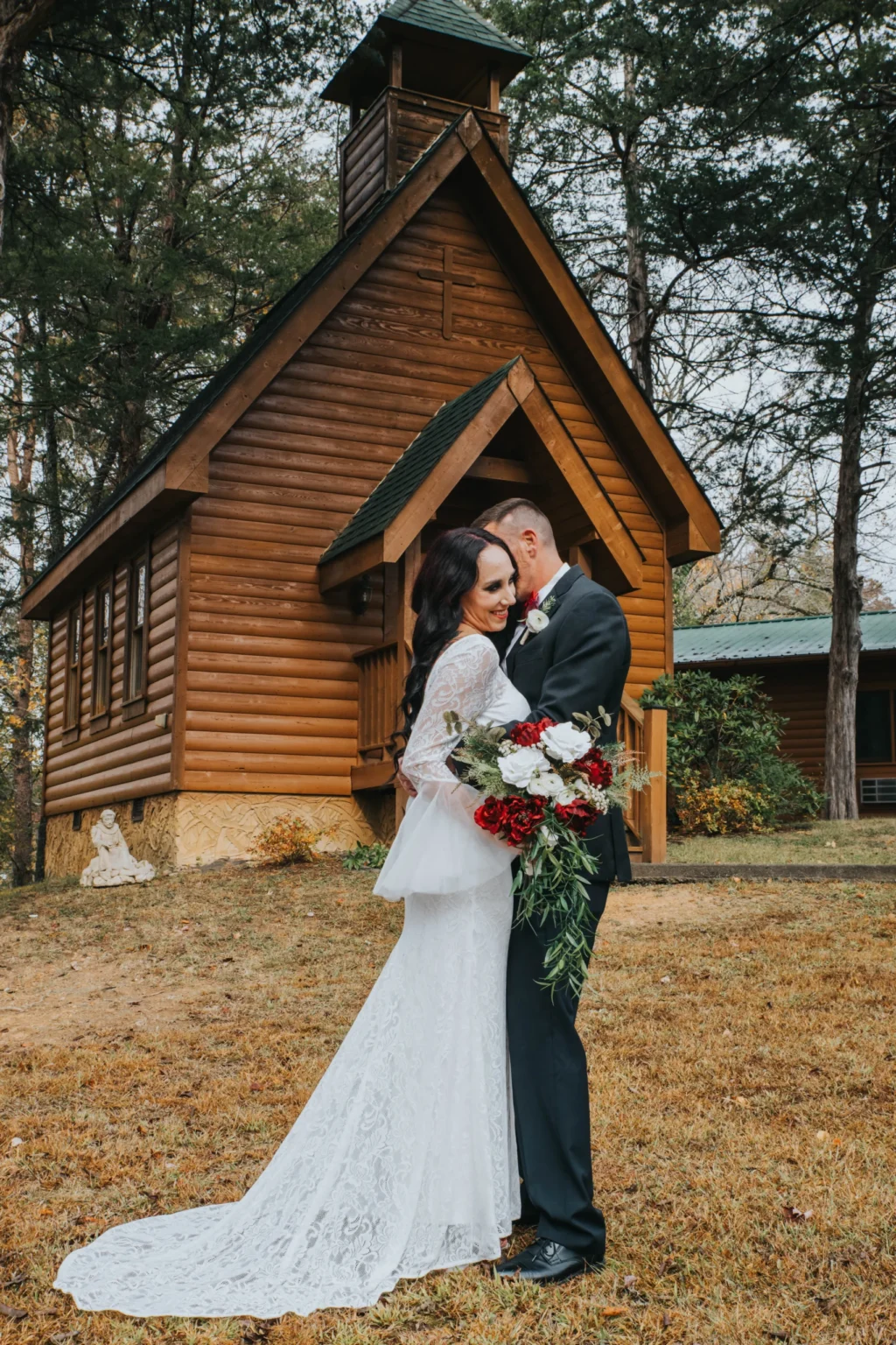 Elopement Wedding A bride in a long, white lace wedding dress and a groom in a black suit with a red rose boutonniere embrace in front of a small, rustic wooden chapel surrounded by trees. The bride holds a bouquet with red and white roses. The scene is outdoors on a slightly overcast day. Elopements Inc