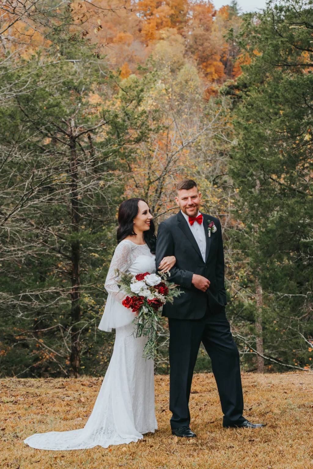 Elopement Wedding A bride in a white lace wedding dress holding a bouquet of red and white flowers stands next to a groom in a black suit with a red bow tie. They are outside with a background of tall trees with autumn foliage. The groom has a hand in his pocket and the couple is smiling as they walk together. Elopements Inc