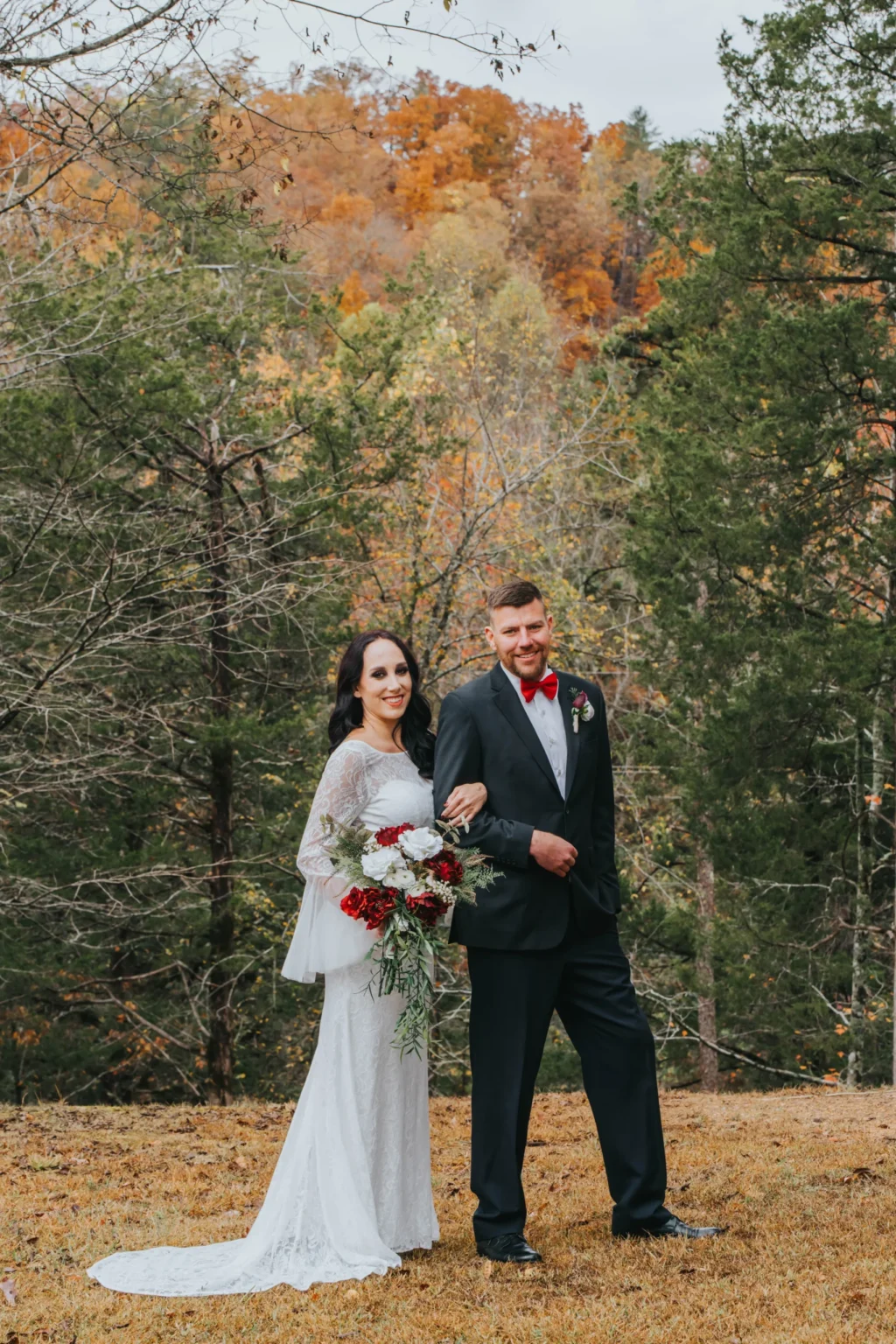 Elopement Wedding A bride in a long white lace dress holds a red and white floral bouquet, smiling, while linking arms with a groom in a black suit with a red bow tie. They stand on grass with autumn trees in the background, creating a colorful and vibrant outdoor setting. Elopements Inc