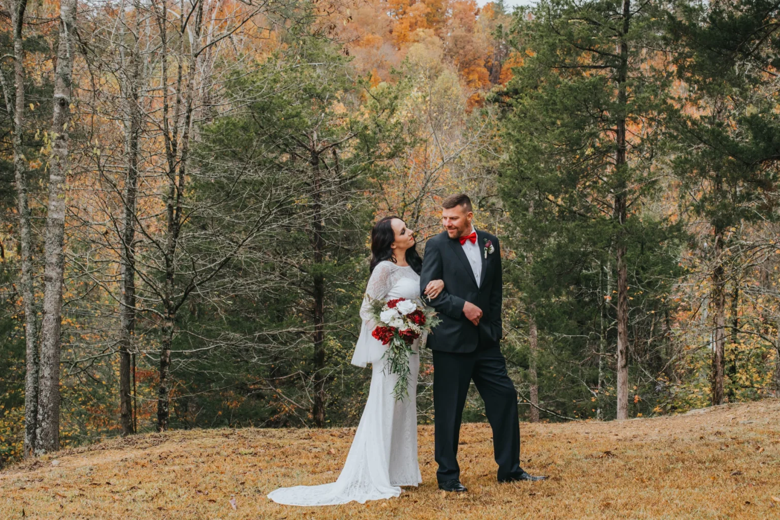 Elopement Wedding A bride and groom stand outdoors in front of an autumn forest backdrop. The bride is in a long white dress with lace sleeves, holding a bouquet of red and white flowers. The groom is in a black suit with a red bow tie. They gaze into each other's eyes, smiling warmly. Fallen leaves cover the ground. Elopements Inc